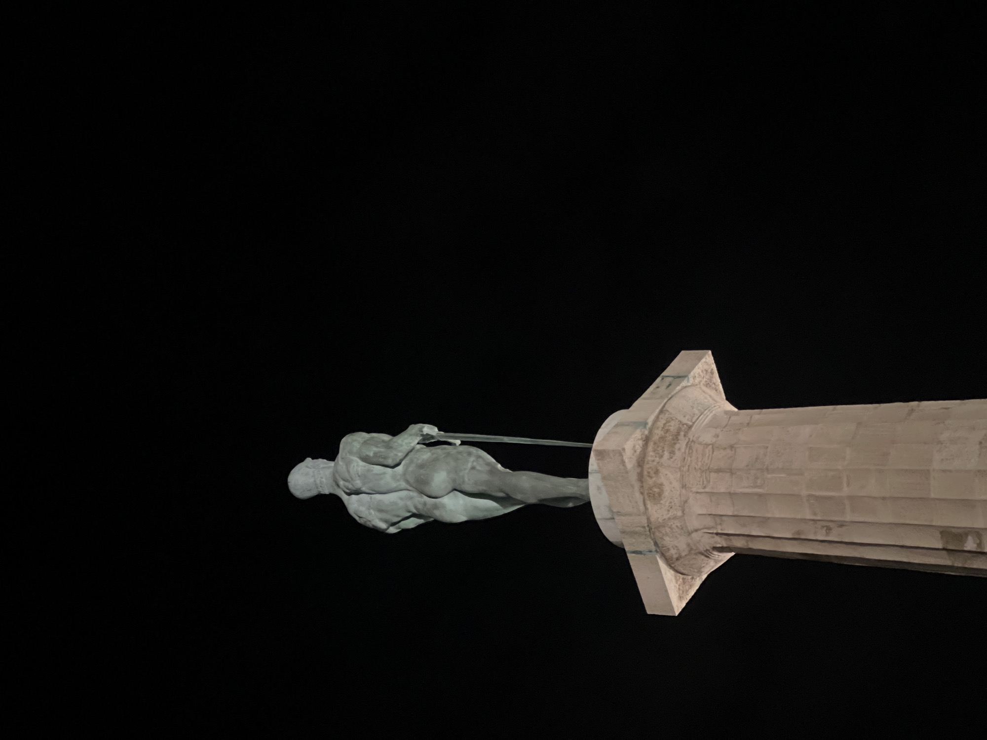 The image shows a statue atop a column against a black background. The statue is a nude male figure holding a sword. The column is light-colored and made of stacked blocks. The location is likely Belgrade, Serbia, and it is probably the Victor monument.