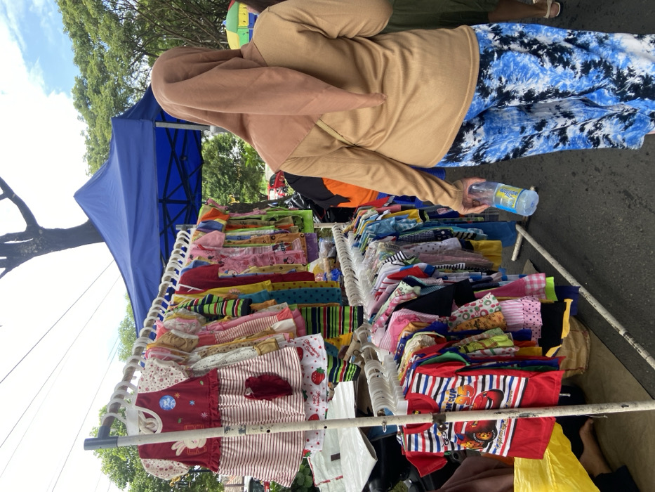 The scene is a street market in Kecamatan Sidoarjo, Indonesia (latitude: -7.441, longitude: 112.705). A clothing rack is loaded with colorful clothes, mainly t-shirts and dresses. A person dressed in brown and a headscarf holds a clear bottle of water in their right hand. The person is walking past the clothing rack and looking toward the right side of the frame. The rack is under a blue canopy tent, and there are trees and other market stalls behind the person.