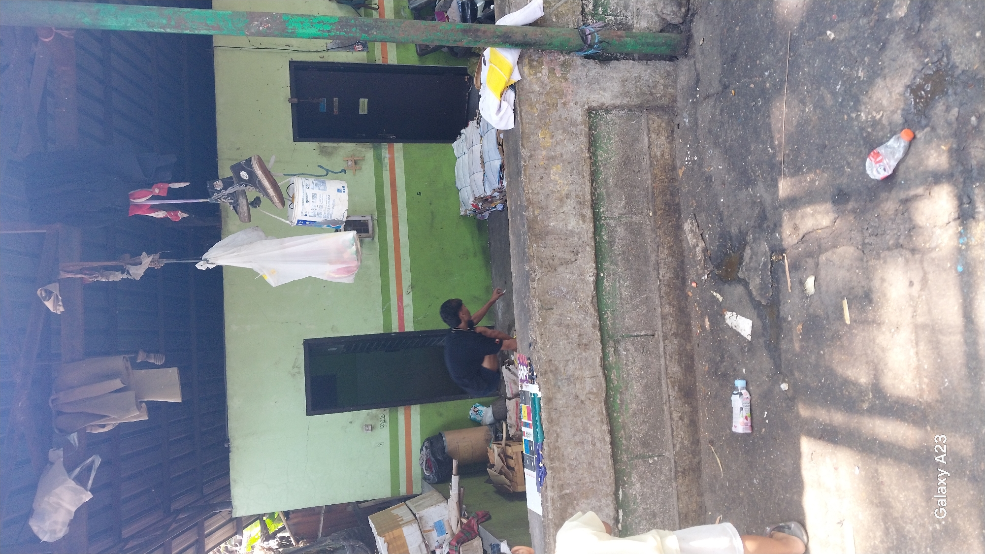 The image depicts an outdoor scene, likely in an alley or an informal settlement area, with a backdrop of a light green wall painted with horizontal stripes of orange and white. Two dark doorways are visible on the wall. A person is crouched down in front of the wall, appearing to be sorting through or handling some items. Above the doorways and extending across the top of the image, a makeshift roof structure with wooden beams is visible, supporting various items hanging from it, including clothes and plastic bags. Shoes are also seen hanging, suspended by their laces. The ground in the foreground is paved and appears somewhat dirty, with scattered debris and a few plastic bottles. The overall impression is of a utilitarian or storage area with evidence of everyday use and some disarray. Based on the "Galaxy A23" watermark in the lower left corner, the image was captured with a Samsung Galaxy A23 smartphone.