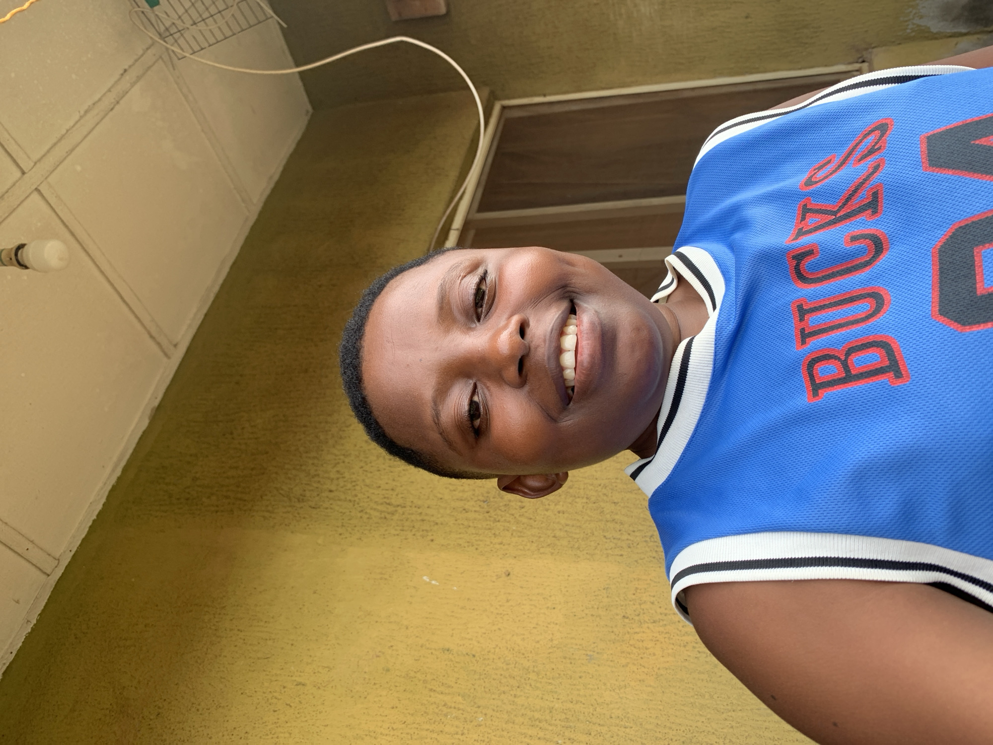 The photograph presents a young individual wearing a blue basketball jersey with the word "BUCKS" visible on the upper right. The individual is smiling and looking directly at the camera. The background consists of a yellow wall. The setting is identified as Ikorodu, Nigeria. There is a white electrical cord visible at the top of the frame.