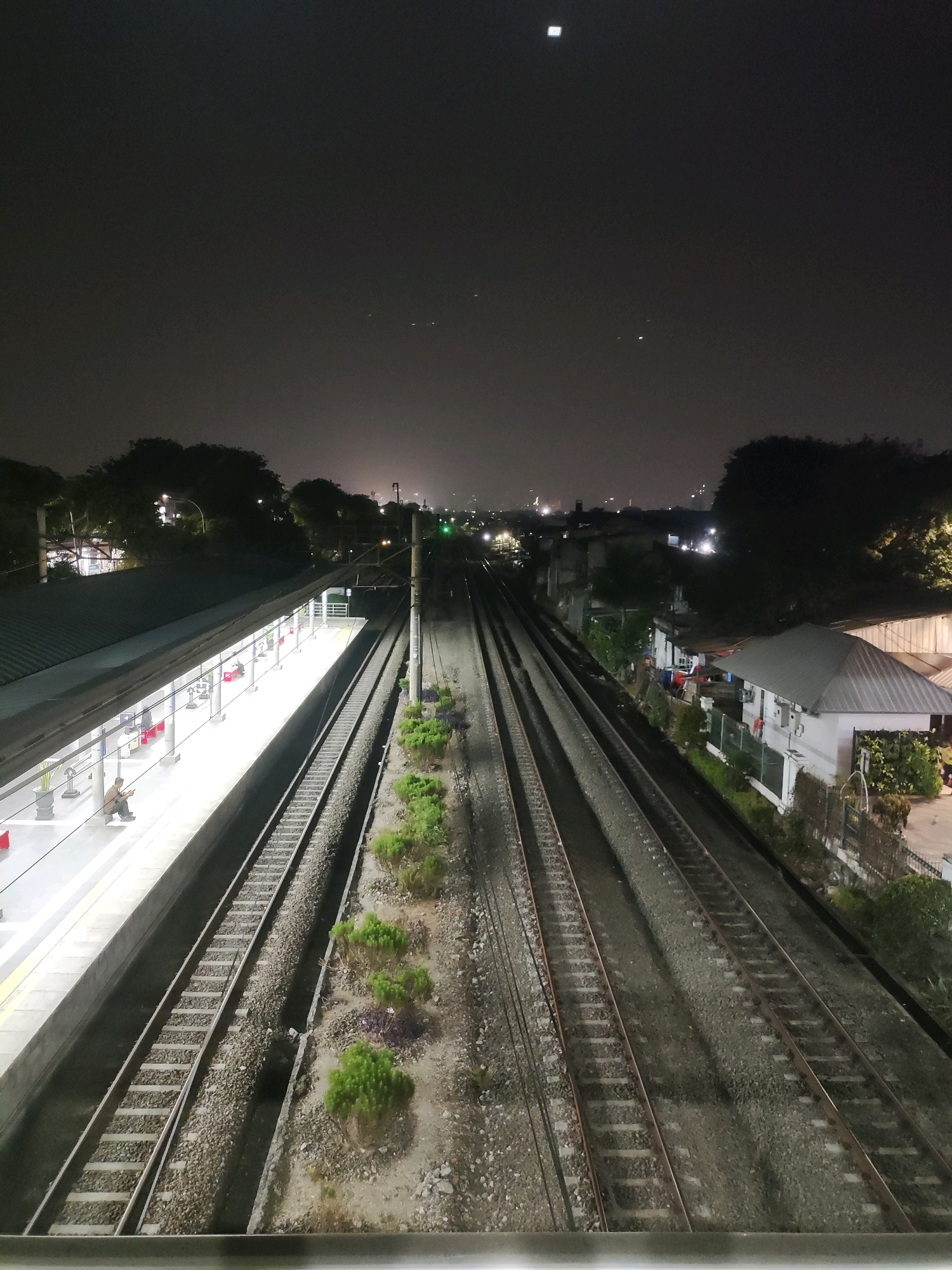 The image depicts a night view of a train station in East Jakarta, Indonesia. In the foreground, a train platform is brightly lit, with a few passengers seated on benches. Train tracks run parallel to the platform, extending into the distance where they merge with other tracks. Buildings and trees line the perimeter of the station, with city lights visible in the background. The sky is dark and cloudy, with a few faint stars visible.