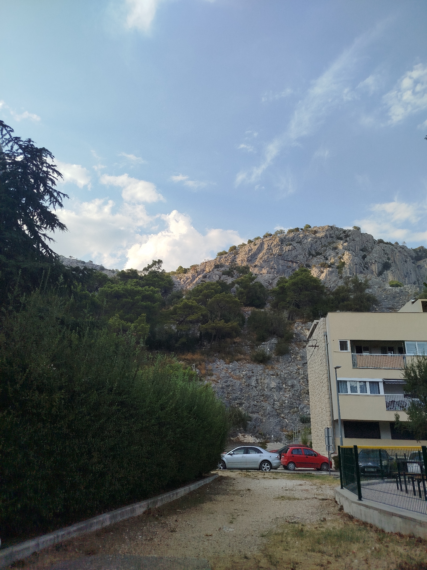 The image depicts a view towards a rocky mountainside and a residential building in Omiš, Croatia. The foreground features a dirt path leading towards a concrete curb and a dense green hedge. To the right of the path, two cars are parked: a silver sedan and a red hatchback. Behind the cars, a multi-story building with light-colored brickwork and balconies is visible. The building has several windows and a black awning over one of them. A black fence with a yellow top encloses an area to the right of the building. The mid-ground is dominated by a steep, rocky slope covered with sparse vegetation, including trees and shrubs. The background is a wide expanse of sky with scattered white clouds. The lighting suggests it is daytime, possibly late afternoon, with the sun casting shadows on the mountain face. The scene appears to be a residential area situated at the base of a natural landscape.