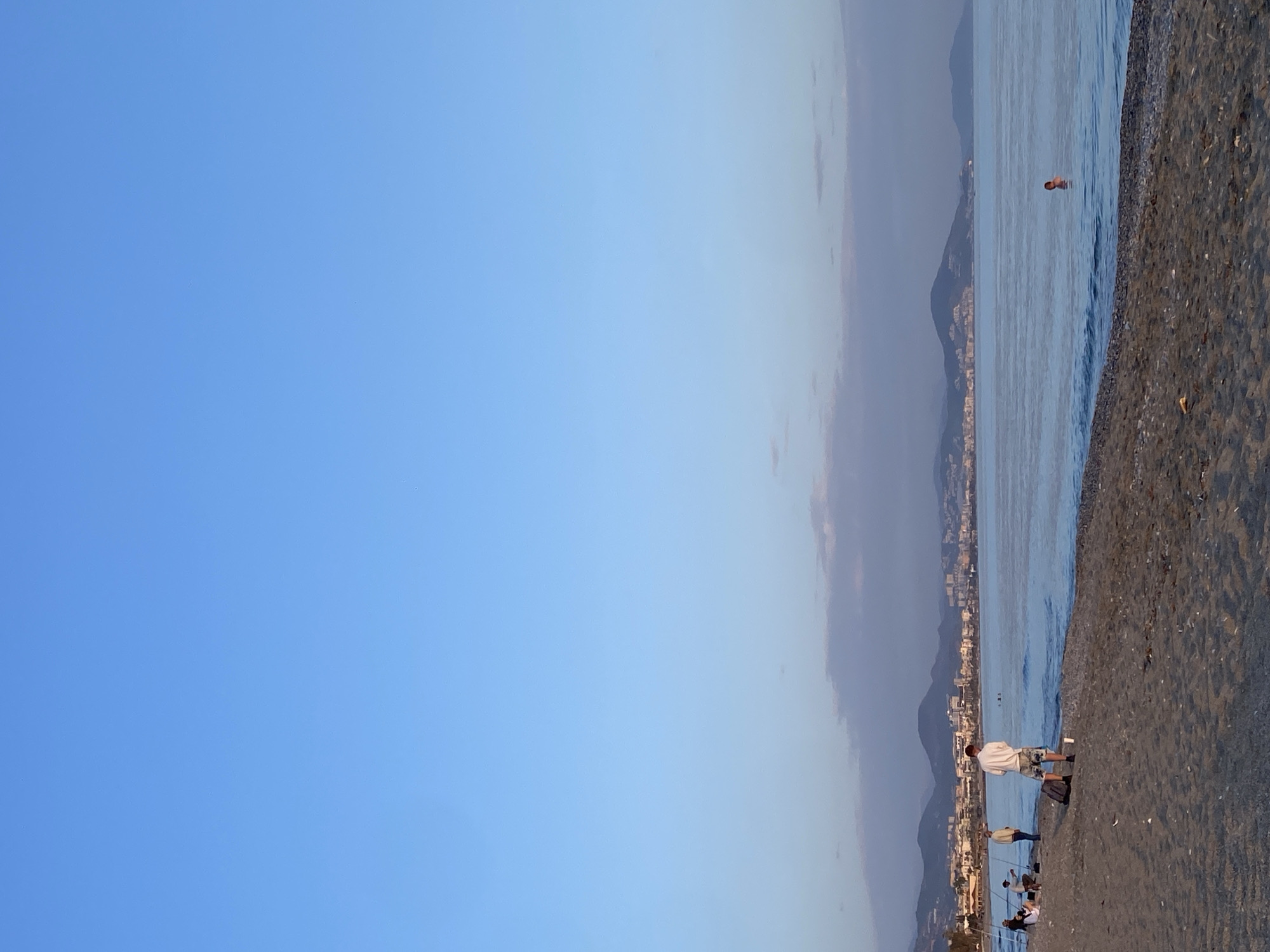 The image captures a coastal scene in Alanya, Turkey. The right side of the frame shows a beach with dark sand and small pebbles. The Mediterranean Sea is to the left of the beach with a person swimming in the distance. In the mid-ground, there are a few people along the shoreline, some possibly fishing with fishing rods. Buildings and mountains form the backdrop. A clear blue sky fills the left side and upper portion of the image.
