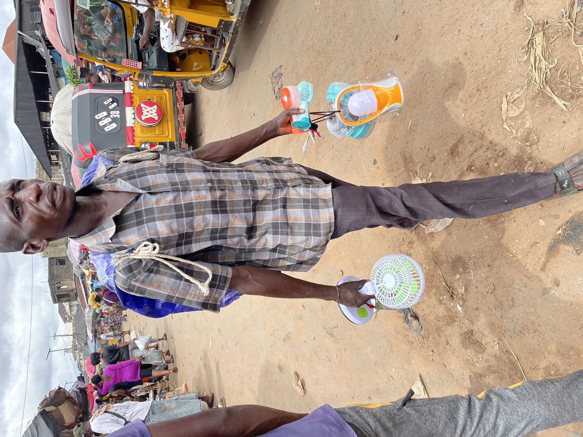 A man walks forward on a dirt road, carrying a bundle on his back and several handheld fans in his right hand. He wears a plaid shirt and dark pants. In the background, a yellow tuk-tuk is parked to the right, and a market scene with various stalls and people is visible. The sky is overcast.
