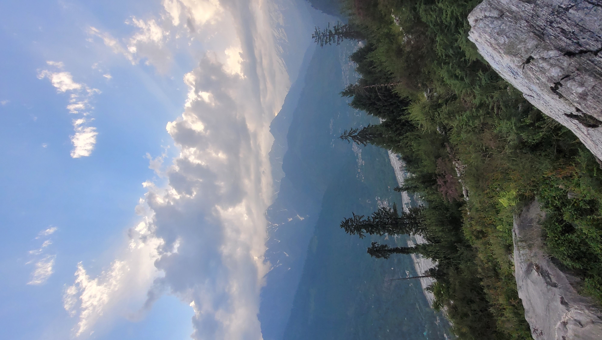 The media file depicts a scenic mountain landscape in Vashisht, India. The image is oriented vertically, showcasing a dramatic view from a rocky vantage point. In the foreground, a large, weathered rock formation dominates the right side of the frame, with its textured surface and cracked details clearly visible. Beyond the rock, a dense forest of evergreen trees stretches out, with their dark green foliage creating a lush carpet.
The mid-ground reveals a valley with a winding river or road, dotted with scattered buildings that suggest a settlement. Further in the distance, majestic mountains rise, their peaks partially obscured by layers of atmospheric haze and soft clouds. Some of the higher mountain slopes show patches of snow, hinting at their elevation and the cooler temperatures at higher altitudes.
The sky above is a vibrant blue, punctuated by a mix of fluffy white clouds and darker, more dramatic formations. Sunlight filters through the clouds, casting rays that illuminate the sky and create a sense of depth. The time of day appears to be late afternoon or early evening, given the soft lighting and the way the light interacts with the clouds. The overall mood is serene and awe-inspiring, highlighting the natural beauty of the Himalayan region. There is no visible text in the image.