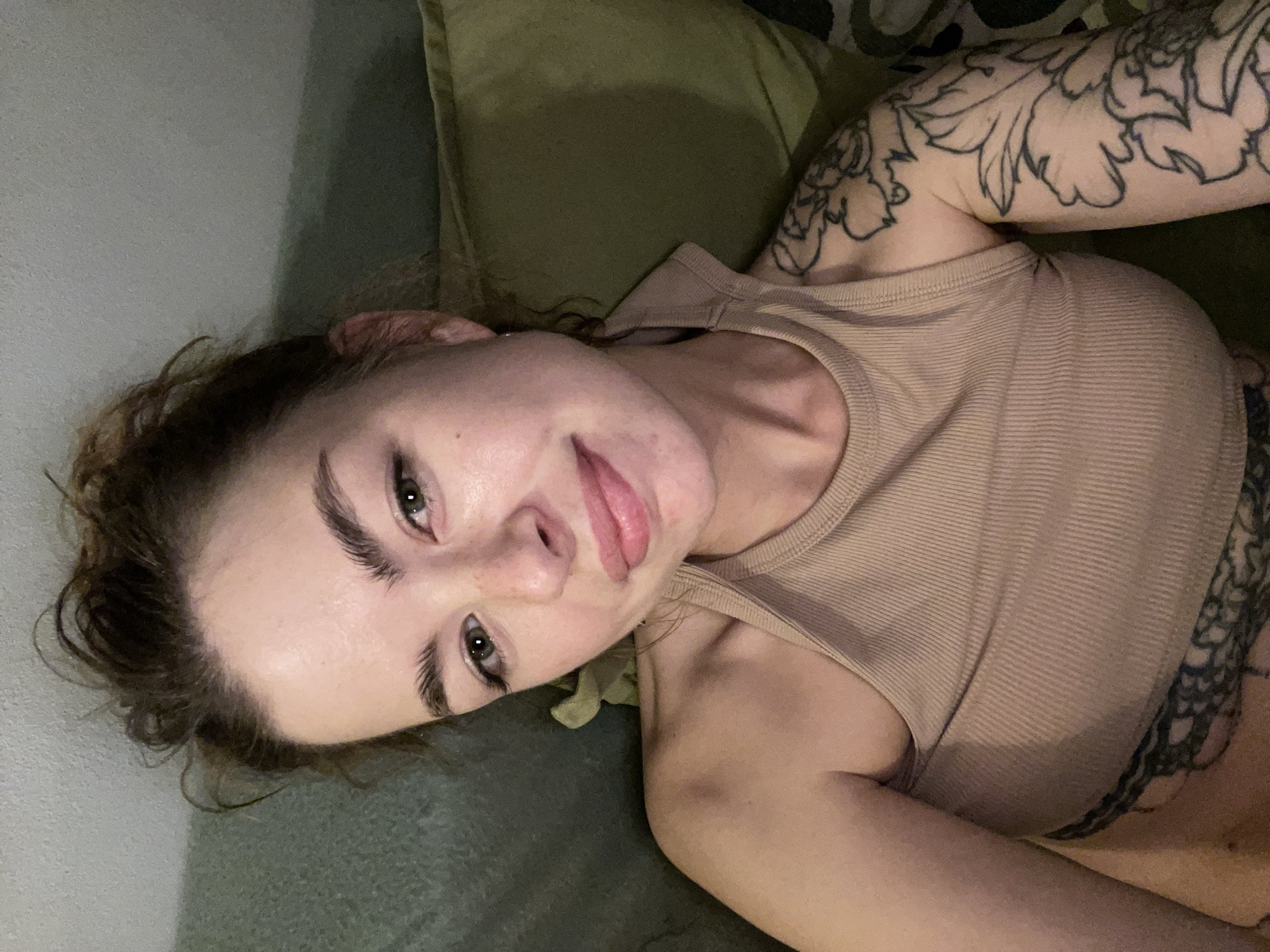 A person is lying on a green couch or cushion. The person has fair skin, brown hair pulled back, and full eyebrows. They are wearing a tan ribbed tank top. Tattoos are visible on the upper arms and side. The location and city cannot be determined from the image.