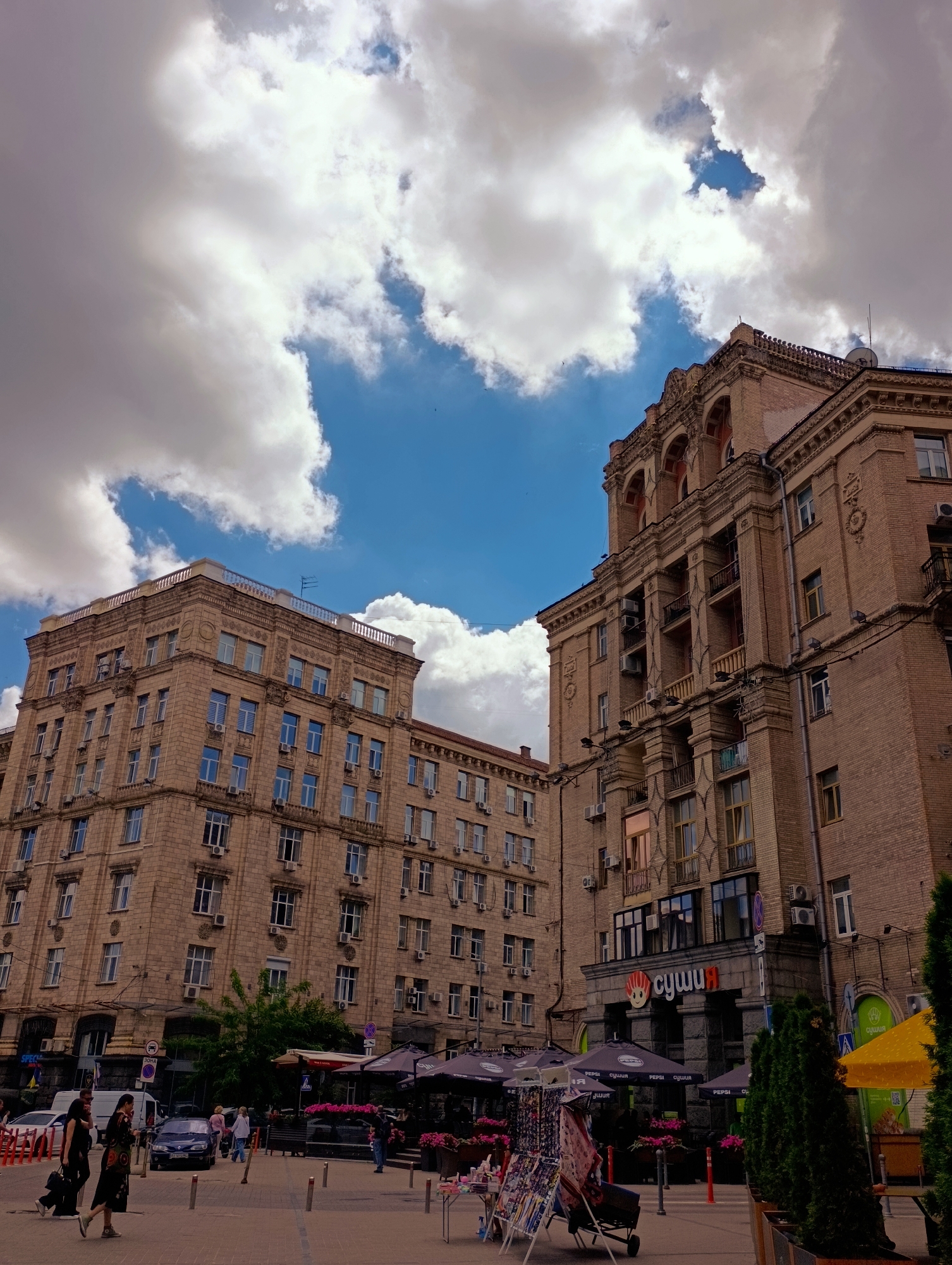 This is a street-level, slightly low-angle shot of a commercial district in Kyiv, Ukraine, on a cloudy day. The foreground features a pedestrian area with several people walking and standing. Two women in dark clothing are walking on the left side of the frame, with one carrying a black handbag. To their right, a dark blue car is parked. In the middle ground, there are several outdoor seating areas with umbrellas, likely for cafes or restaurants. One umbrella is branded with "PEPSI". In the background, two large, multi-story brick buildings dominate the scene. The building on the right has a distinctive facade with arched windows and ornate detailing. A sign with Cyrillic text, "СушиЯ", is visible on this building, indicating a sushi restaurant. The building on the left is also substantial, with numerous windows. The sky above is filled with dramatic, large cumulus clouds, with patches of blue sky showing through. The overall impression is a bustling urban environment under a dynamic sky.