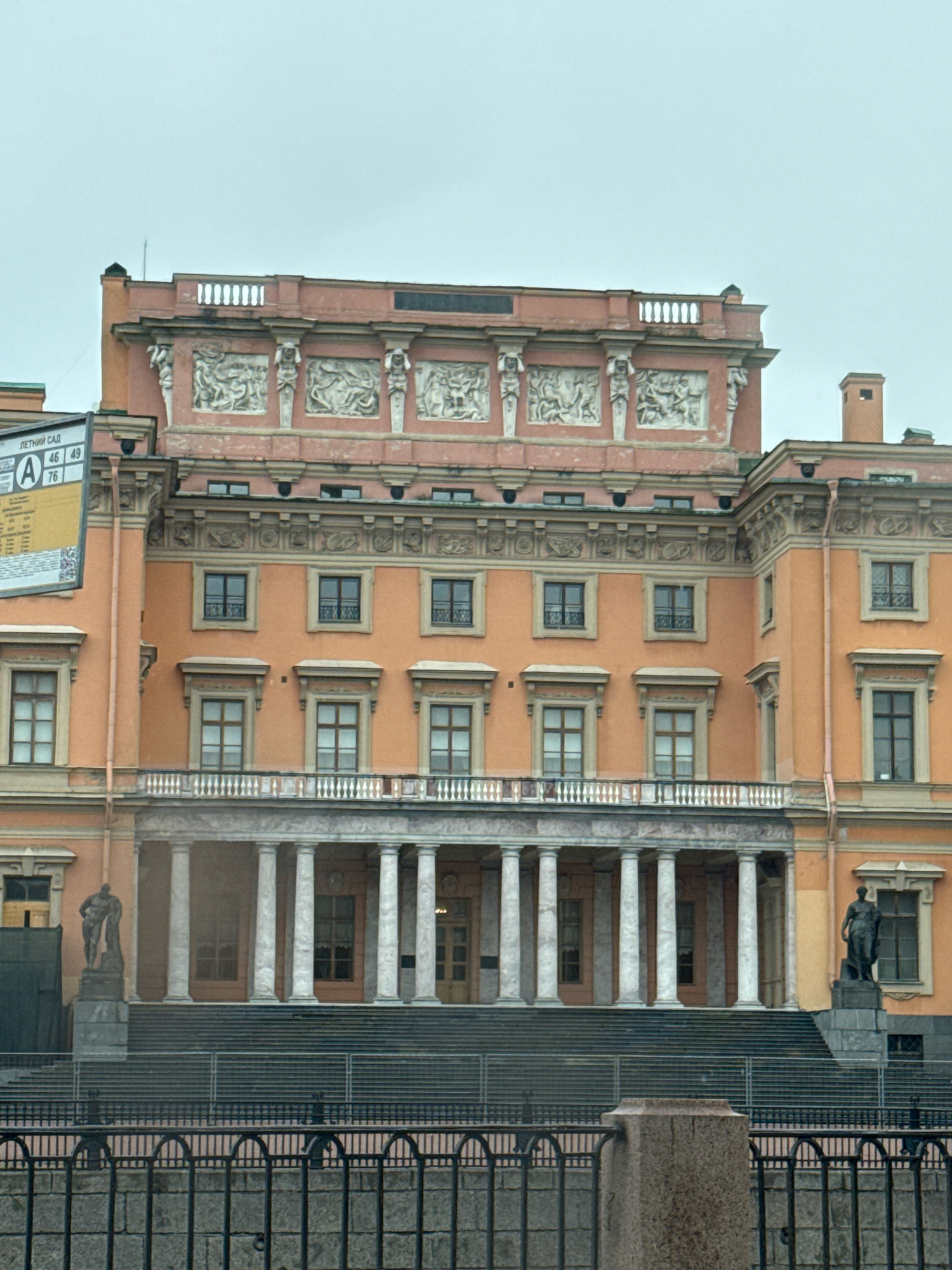 The image depicts the Mikhailovsky Castle in Saint Petersburg, Russia. The castle, with its distinctive salmon-pink facade, is a prominent architectural landmark. The building features classical elements such as columns and decorative reliefs above the windows. In front of the castle, a wide staircase leads up to a portico supported by marble columns. Two statues are visible: one on the left, near a dark tarp-covered area, and another on the right, standing on a pedestal. A black wrought-iron fence runs along the foreground, with a stone pillar supporting part of it. The sky above is overcast, suggesting a cloudy day. A street sign with Russian text and numbers is partially visible on the left side of the frame.