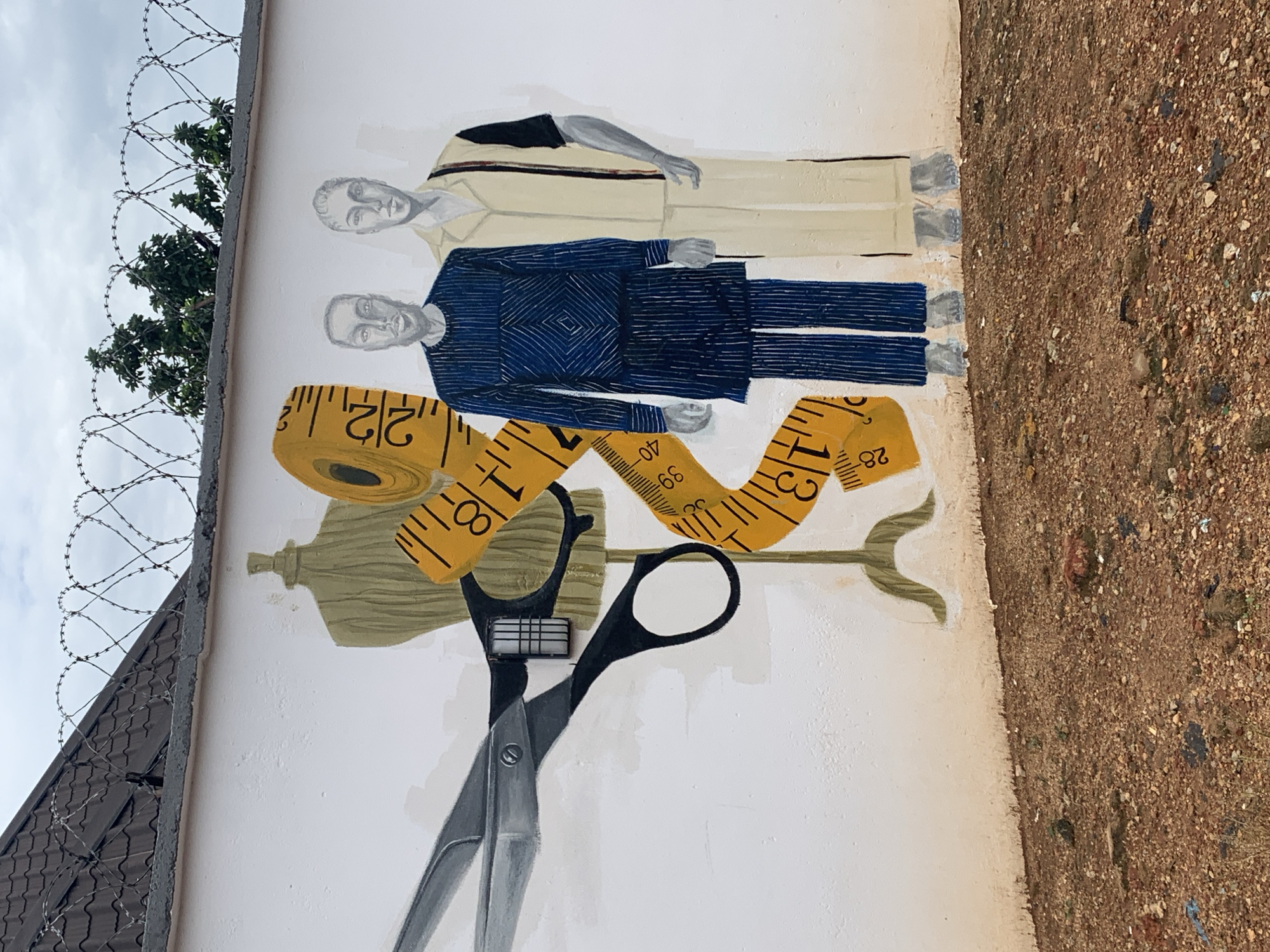A mural on a white concrete wall depicts a tailor's theme, located in Gura Top, Nigeria. On the left, a large pair of scissors is painted, alongside a dressmaker's mannequin in a muted green. A bright yellow measuring tape, marked with black numbers and increments, curves across the wall, wrapping around the mannequin and extending towards the right. To the right of these elements, two figures are painted, appearing as stylized human figures. The figure on the left wears a dark blue garment with vertical stripes and a diamond pattern on the chest. The figure on the right wears a light-colored shirt with black and red trim on the sleeves and a matching pair of light-colored pants. Both figures are depicted with gray skin tones. The wall is situated above a dry, pebbly ground. Barbed wire coils are visible along the top edge of the wall, with green foliage and trees visible behind it. The sky is overcast.