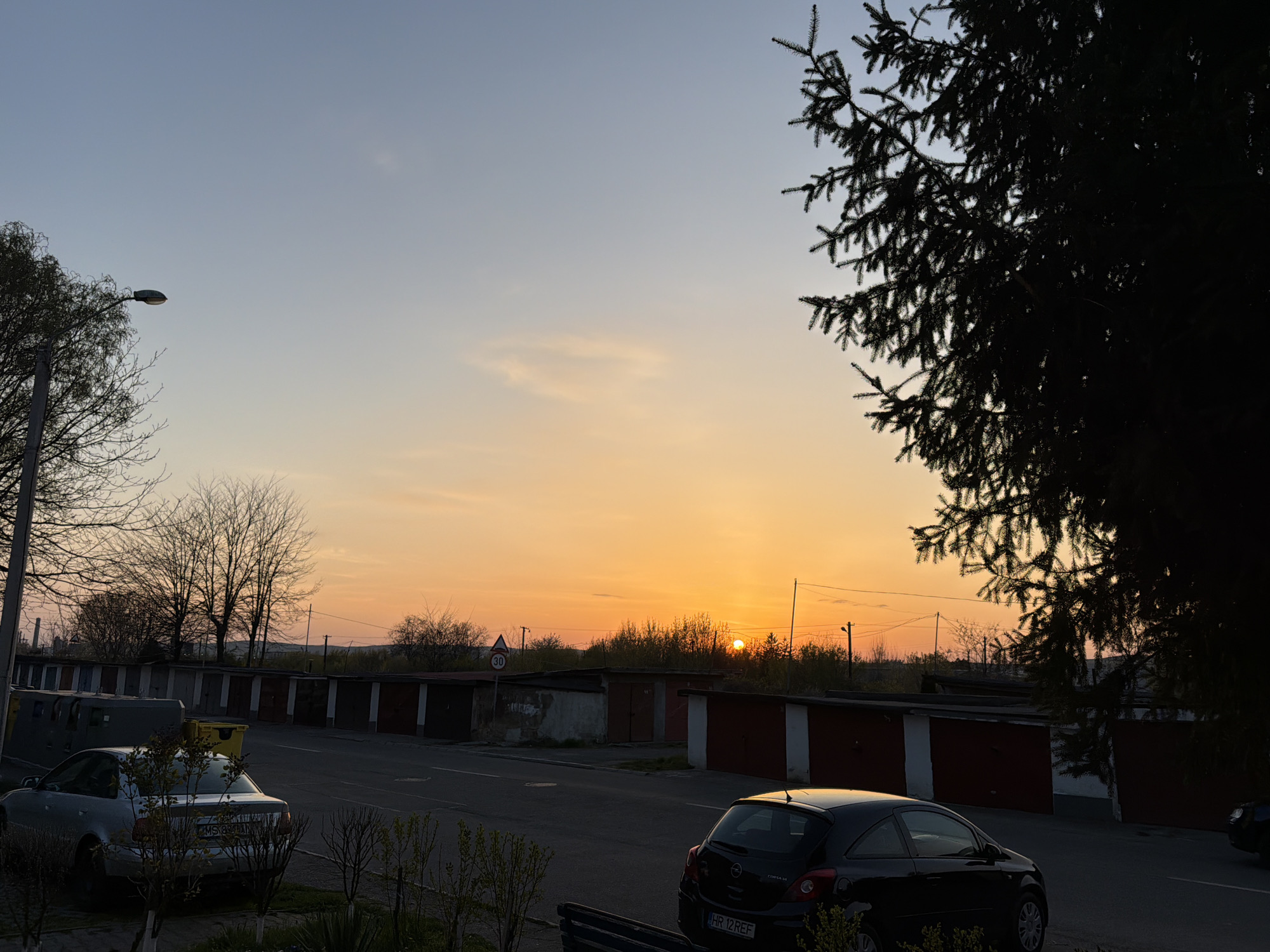 The image shows a street in Târgu Mureș, Romania, lined with a series of garages under a sunset sky. A silver sedan is parked in the foreground on the left, while a black hatchback is parked on the right. The sky is transitioning from blue to orange as the sun sets behind the garages. There is a 30 km/h speed limit sign on the street. A few bare trees and a large evergreen are visible on the edges of the image. A streetlight is present on the left side of the street.