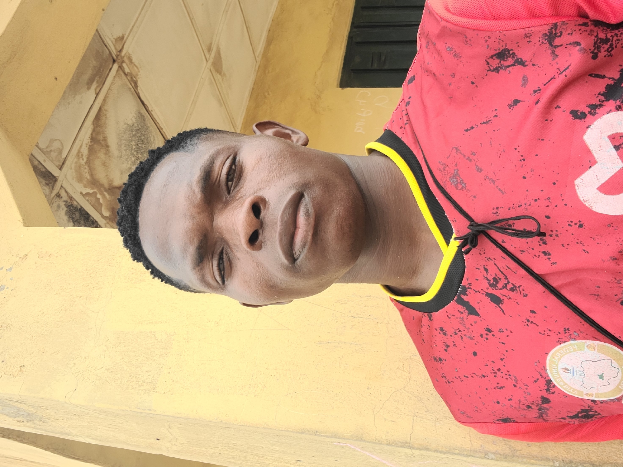 The media file shows a close-up portrait of a young Black person with dark skin and short, curly black hair. They are wearing a red t-shirt with black and yellow trim. The t-shirt has a distressed, splattered pattern in black and a circular emblem on the left chest. The person's gaze is directed towards the camera, with a neutral expression.

The background appears to be an exterior wall painted a light yellow, with some visible cracks and water stains. To the upper right, a dark, slatted window or shutter is partially visible. The lighting suggests it is daytime, possibly sunny, given the shadows and the brightness of the scene. The composition is a selfie-style shot, with the camera held at a high angle, looking down at the subject.

The provided location context of Masaka, Nigeria, suggests the setting is likely in this region. No specific activity or event is discernible beyond the portrait itself. The image does not contain any clearly legible text, apart from what might be part of the emblem on the t-shirt, which is too pixelated to read.
