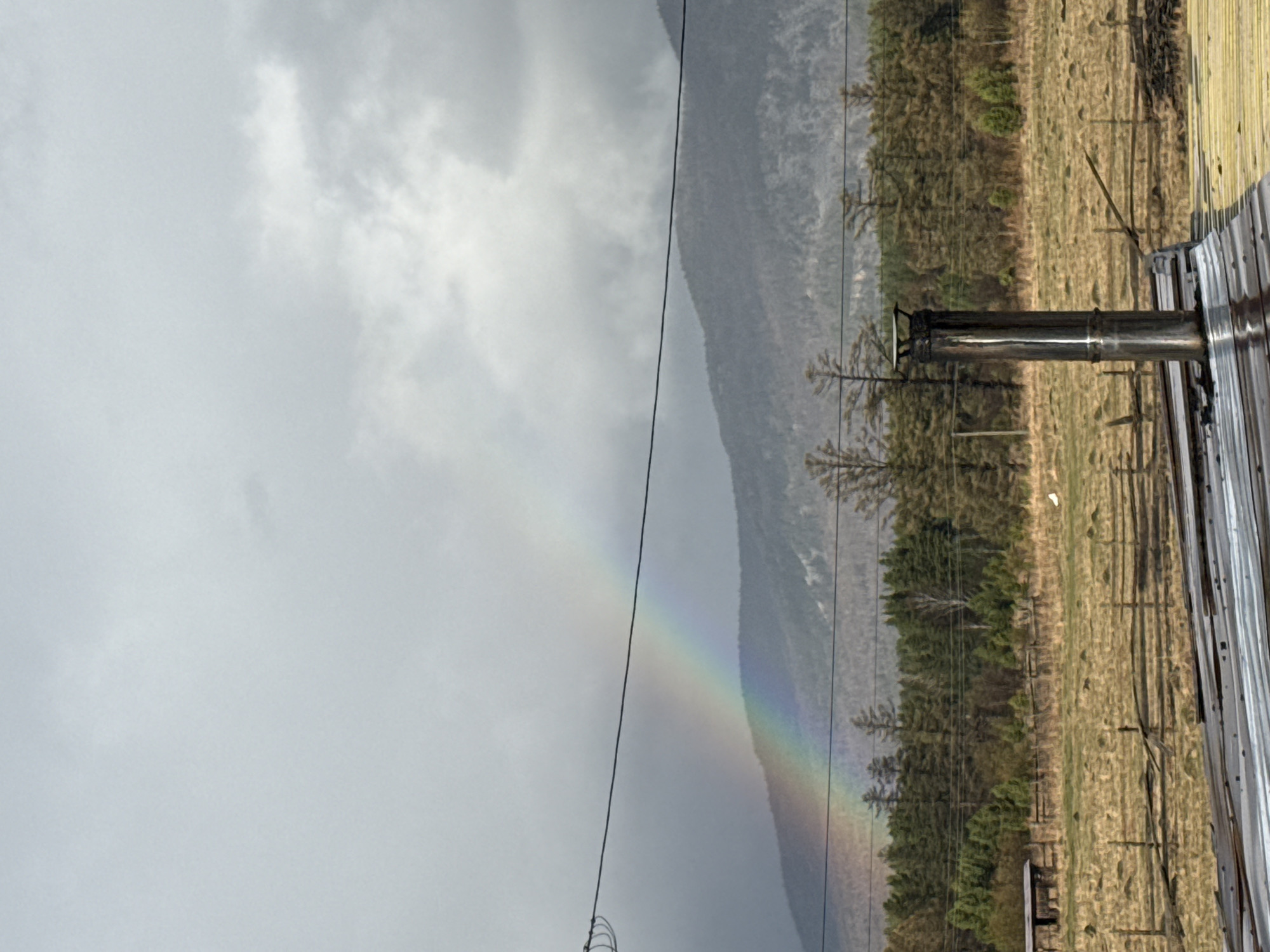 A landscape image depicts a rainbow arcing across the sky, terminating near a forested hillside in the background. The foreground includes a metal chimney extending from a metallic structure or roof. Power lines cut across the scene. The sky is overcast. The location is suggested to be Tyulyuk, Russia, based on the prompt.