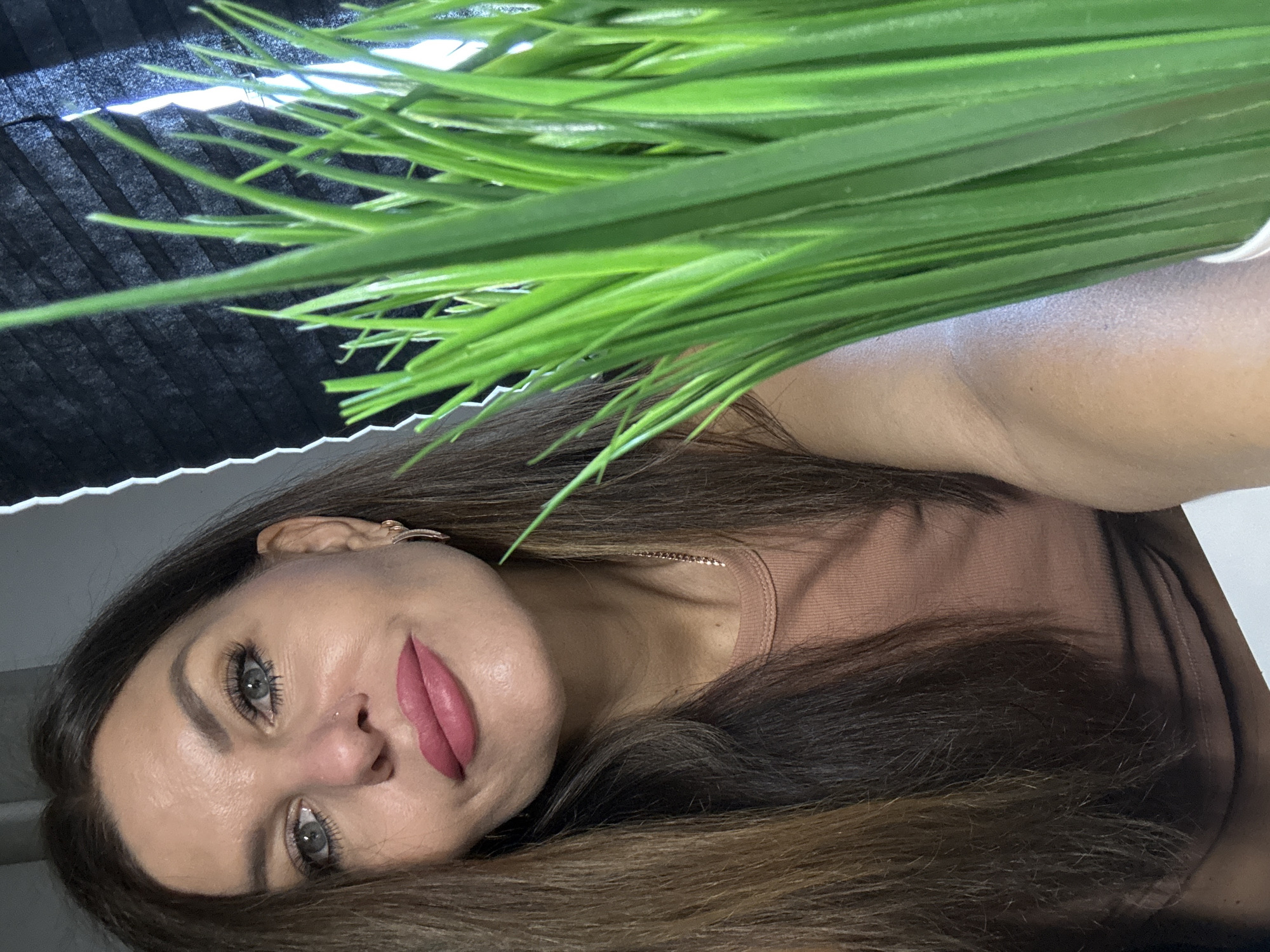 The image shows a close-up of a woman's face, with a potted plant of bright green grass in the foreground, partially obscuring her right side. The woman has long, brown hair, blue eyes, and is wearing pink lipstick and a salmon-colored shirt. She is looking towards the camera with a slight smile. The background consists of a dark, textured surface, possibly a blind, with a sliver of white light visible on the right. The overall lighting is soft, and the focus is on the woman's face and the grass. The image does not contain any contextual information to place it in Chaikovsky, Russia, or any other specific city.