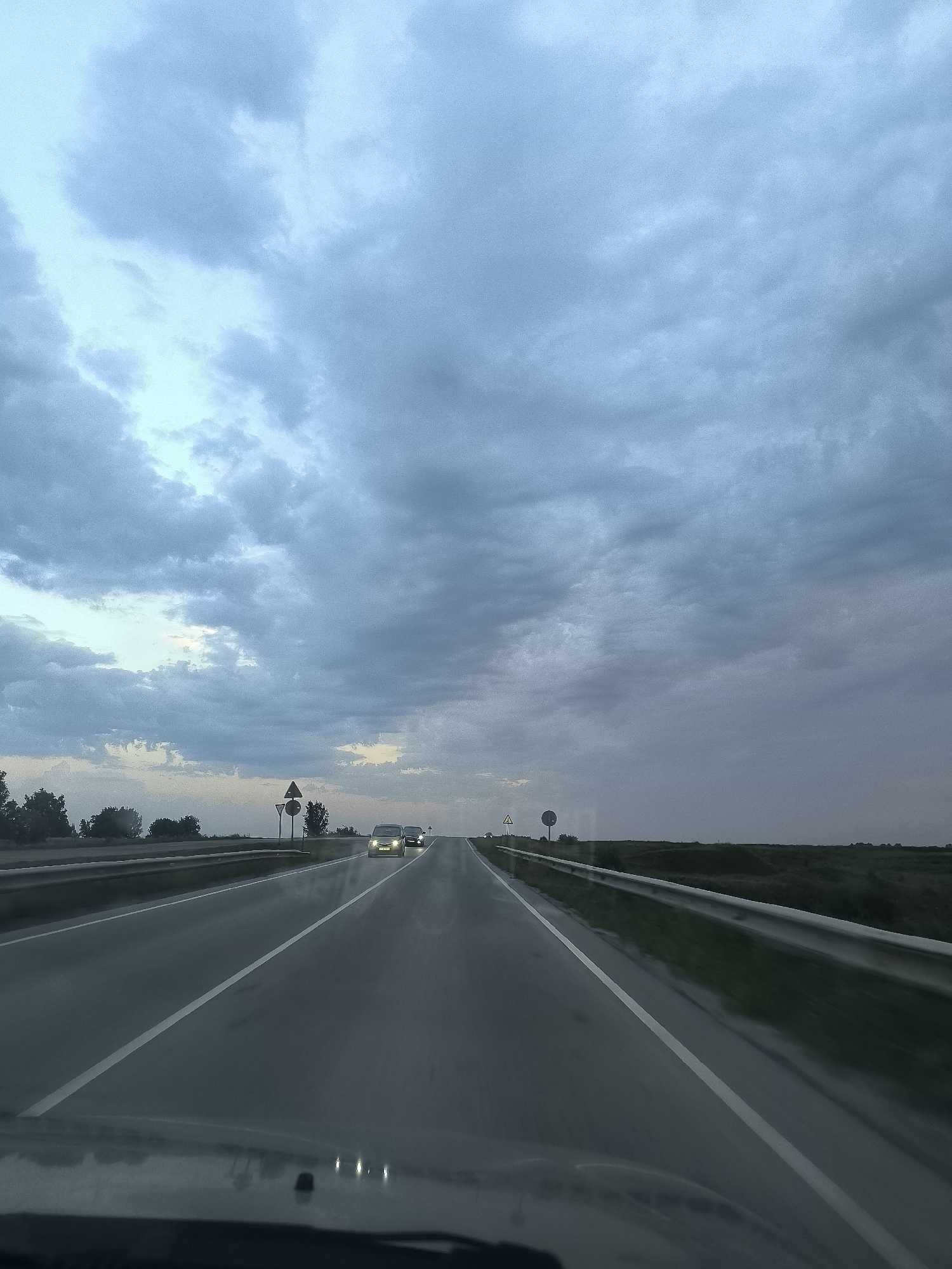 The image depicts a view from the perspective of a vehicle traveling on a multi-lane highway in the vicinity of Krasny Yar, Russia. The sky is overcast with heavy, gray clouds, suggesting a potentially stormy or twilight atmospheric condition. The road is paved and wet, with visible white lane markings. Two cars are traveling ahead in the same direction. Roadside elements include a metal guardrail on the right side of the road and trees and vegetation bordering the highway on the left. Several road signs are visible on the left side of the road, including a triangular warning sign and a circular sign. The overall impression is that of a journey on a highway under a dramatic, cloudy sky.