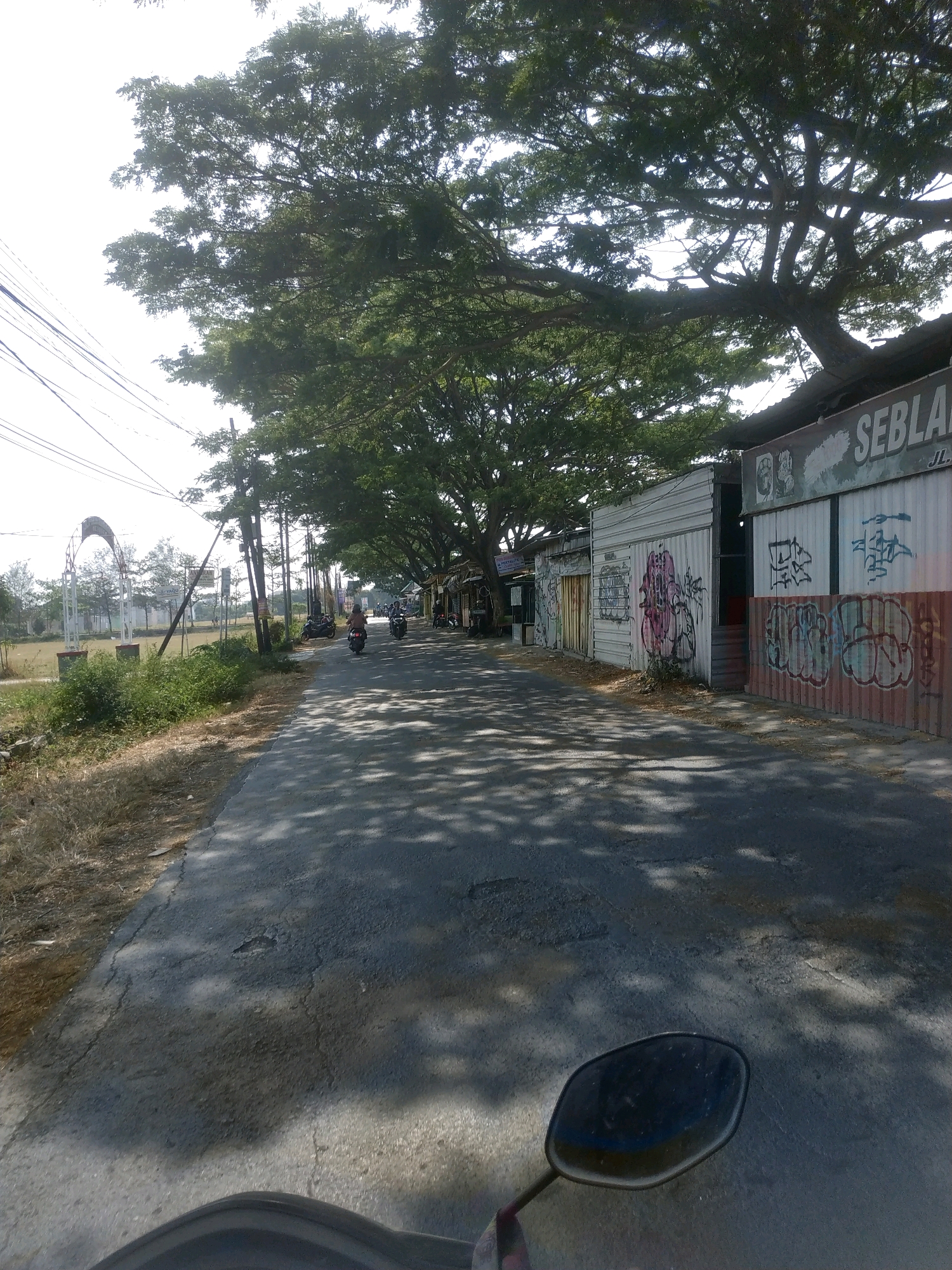 The image shows a street scene in Mejasem Timur, Indonesia. A paved road stretches into the distance, lined with buildings on the right and trees on the left. The buildings on the right appear to be small shops or structures with corrugated metal walls, some decorated with graffiti. A large tree with dense foliage casts shadows across the road. Several motorcycles are visible on the road, with some individuals riding them. In the background, there is an open field with an archway structure. The sky is bright, suggesting a sunny day. A portion of a motorcycle's rearview mirror and handlebars are in the foreground, indicating the image was taken from a moving vehicle.