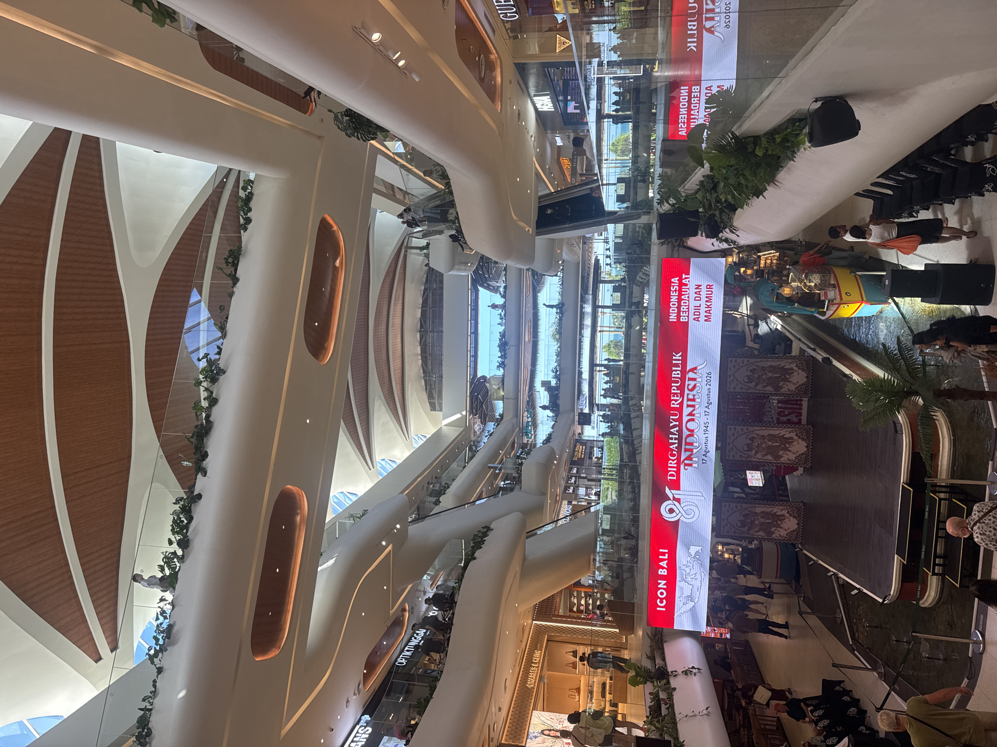 The image captures the interior of a modern, multi-level shopping mall in Denpasar, Indonesia, likely the Beachwalk Shopping Center, known for its organic, flowing architectural design. Sunlight streams in from large open spaces, illuminating the light-colored curved walls and wooden ceiling elements. Lush green plants are strategically placed along the balconies and walkways, adding to the natural ambiance.
In the foreground, a large LED screen displays a festive message for "DIRGAHAYU REPUBLIK INDONESIA" (Long Live the Republic of Indonesia), with the dates "17 Agustus 1945 - 17 Agustus 2026" indicating a celebration of Indonesia's independence. The screen also features the text "ICON BALI" and "INDONESIA BERDAULAT ADIL DAN MAKMUR". Below the screen, a wooden stage is set up, with traditional Balinese-style artwork visible on a backdrop. A small, colorful boat-like structure with a vendor selling drinks is positioned near the stage, drawing attention.
People are seen browsing shops, walking on escalators and walkways, and gathering around the stage area. Shops like "CHARLES & KEITH" and "GUESS" are visible, along with other branded storefronts. The mall's layout features multiple interconnected levels, creating a sense of depth and spaciousness. Reflections can be seen in the glass balustrades and shop windows. The overall impression is one of a vibrant and contemporary retail and entertainment space.