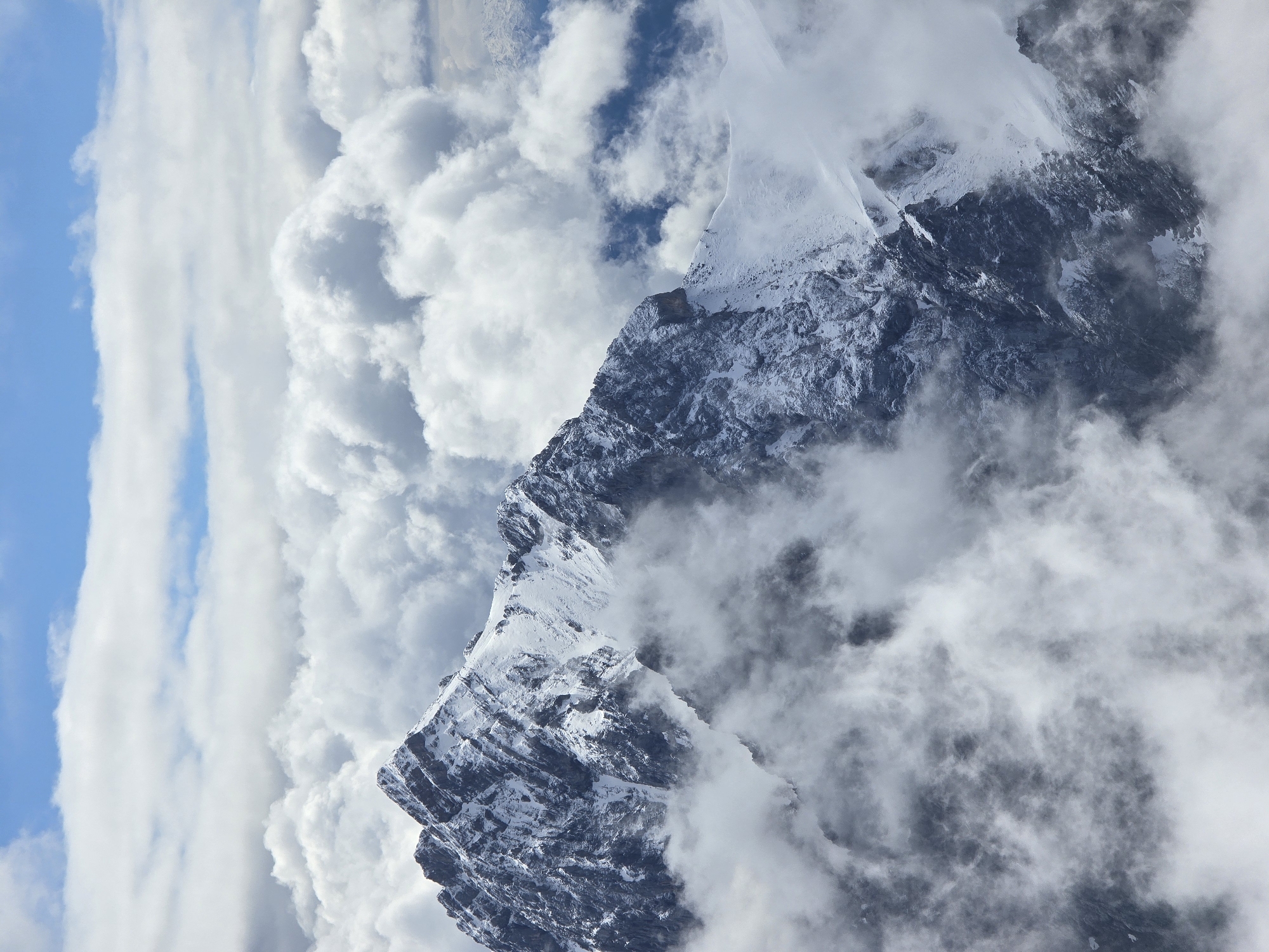 The image shows a majestic snow-capped mountain peak emerging from a sea of clouds. The mountain's rugged, rocky slopes are dusted with snow, creating a stark contrast between the dark rock and the white snow. Wisps of clouds drift around the peak, partially obscuring its form and adding a sense of depth and mystery to the scene. In the background, patches of blue sky are visible, peeking through the clouds. The overall impression is one of dramatic, natural beauty, with the mountain appearing as a solitary sentinel above the clouds. This scene is reminiscent of the Zugspitze, Germany's highest peak, located near Garmisch-Partenkirchen.