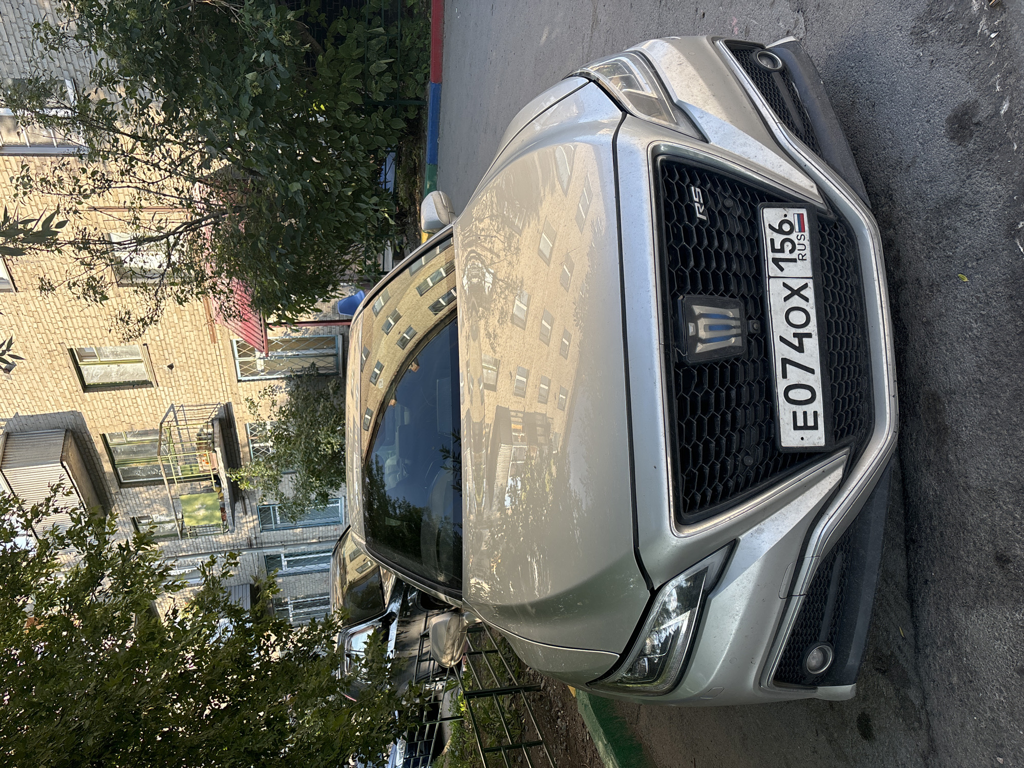 A light-colored sedan is parked in front of a brick apartment building. The car is the main subject, positioned centrally in the frame. Its front grille features a honeycomb pattern and a logo. A license plate is visible on the front bumper, displaying "E074OX 156 RUS". Reflections of the apartment building are visible on the car's hood and windshield. In the background, trees with green foliage partially obscure the building. The building itself is made of light-colored bricks and has multiple windows and balconies. A black SUV is parked behind the sedan, partially visible. The scene appears to be a residential courtyard or parking area in a city.