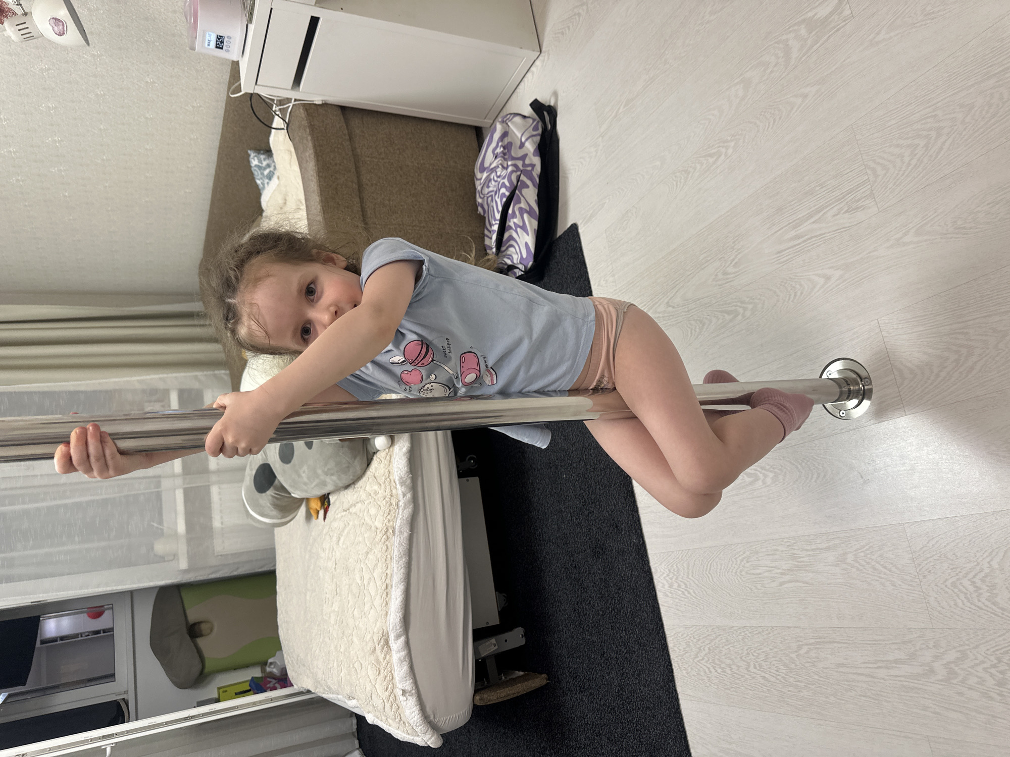 A child is holding onto a horizontal metal bar in a room in Tomilino, Russia. The child wears a light blue t-shirt and pink underwear. The bar is mounted to the wall, and the child is positioned perpendicular to it. The room contains a couch, a white cabinet, a black rug, a bed with a white quilted cover, and a window with blinds. A stuffed animal is visible on the bed. A clothes rack and open shelving unit are partially visible beneath the window.