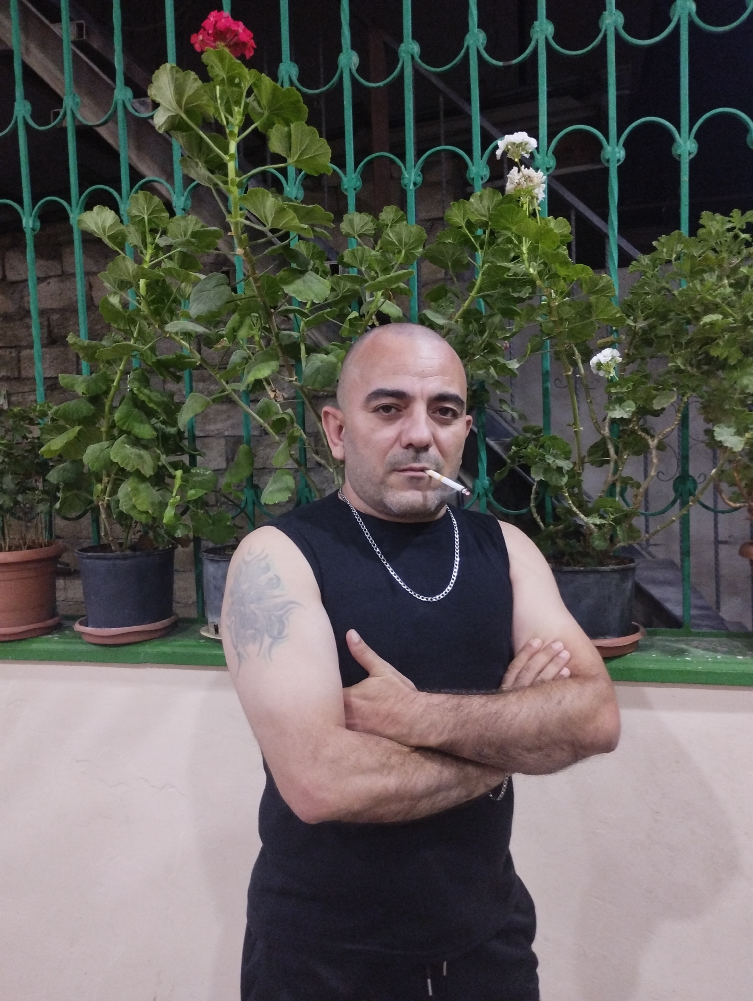 A middle-aged man with a shaved head and a tattoo on his left arm stands with his arms crossed, smoking a cigarette. He is wearing a black sleeveless shirt and a silver chain. Behind him, several potted plants are visible in front of a green metal grate. The scene appears to be an outdoor setting, possibly a balcony or patio, in the city of Bilajari, Azerbaijan.