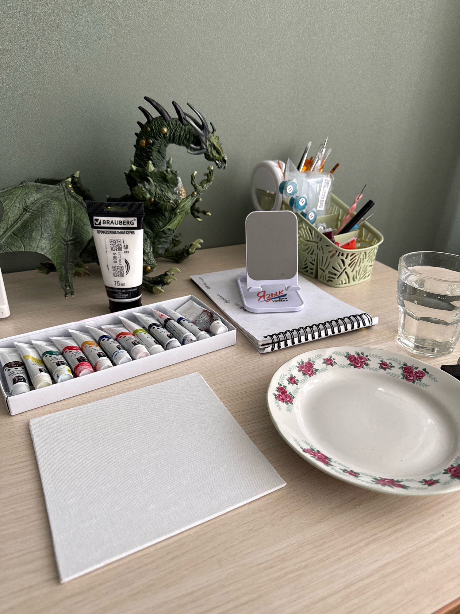 The image depicts a still life of art supplies arranged on a wooden desk. A green dragon figurine is positioned prominently on the left side. In front of the figurine, a tube of white paint by BRAUBERG is visible, along with a box of small paint tubes in various colors. A white canvas is placed in the foreground, to the left. To the right of the canvas, there is a white plate with a floral pattern. A glass of water and a spiral-bound notebook with a phone stand are also present on the desk. The background consists of a textured wall. The scene is an indoor setting, likely an artist's workspace. There is no discernible connection to the city of Cheboksary, Russia, or any specific location within it.