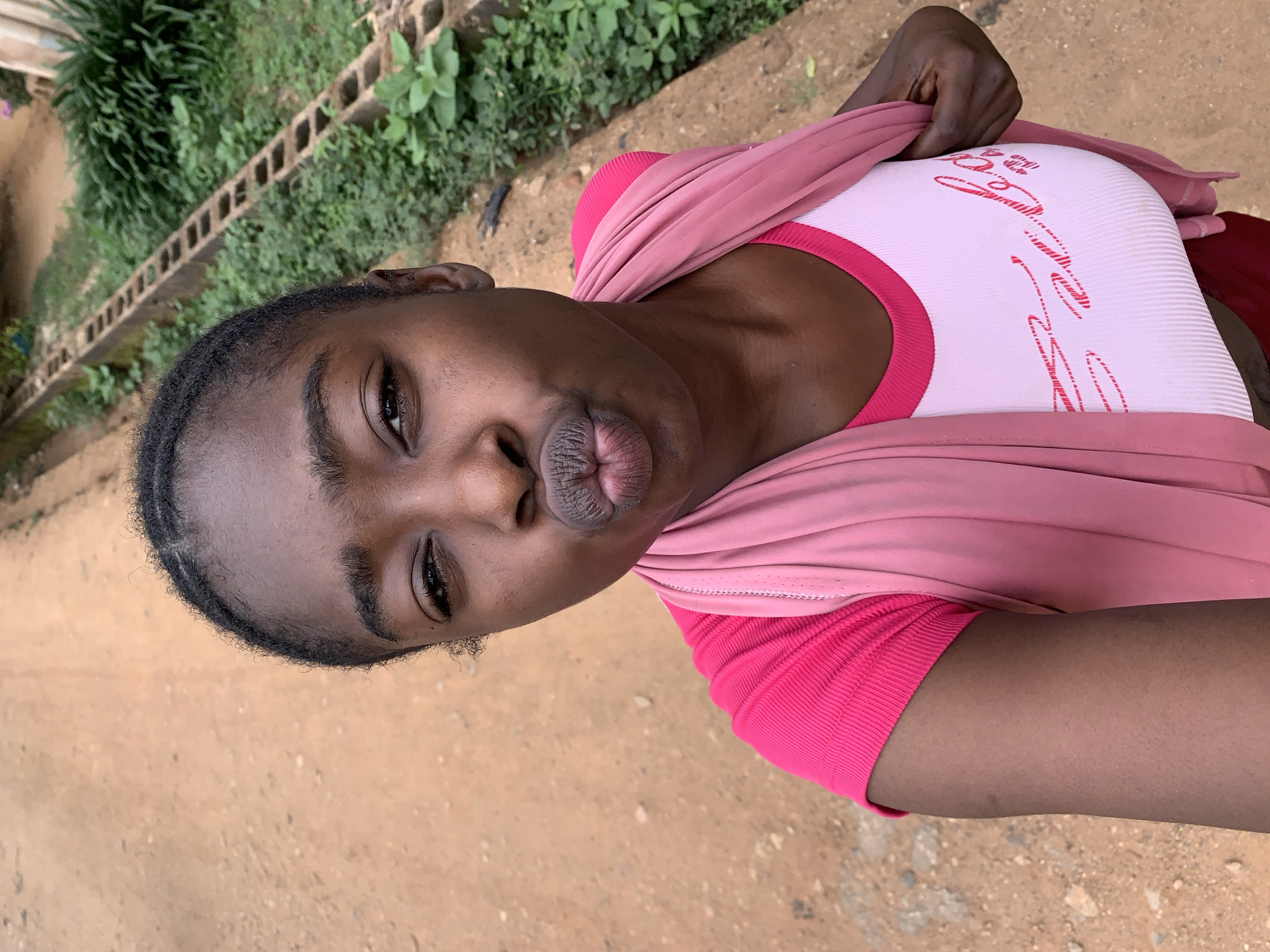 A dark-skinned person with a pout poses for a picture outside. The person is wearing a white tank top with pink trim and graphic, a pink top with short sleeves, and a pink shawl. The person is on a brown dirt surface, possibly a road. In the background there is green vegetation and a cinder block wall. The photograph was taken in a natural outdoor environment, likely in Rafin Par, Nigeria.