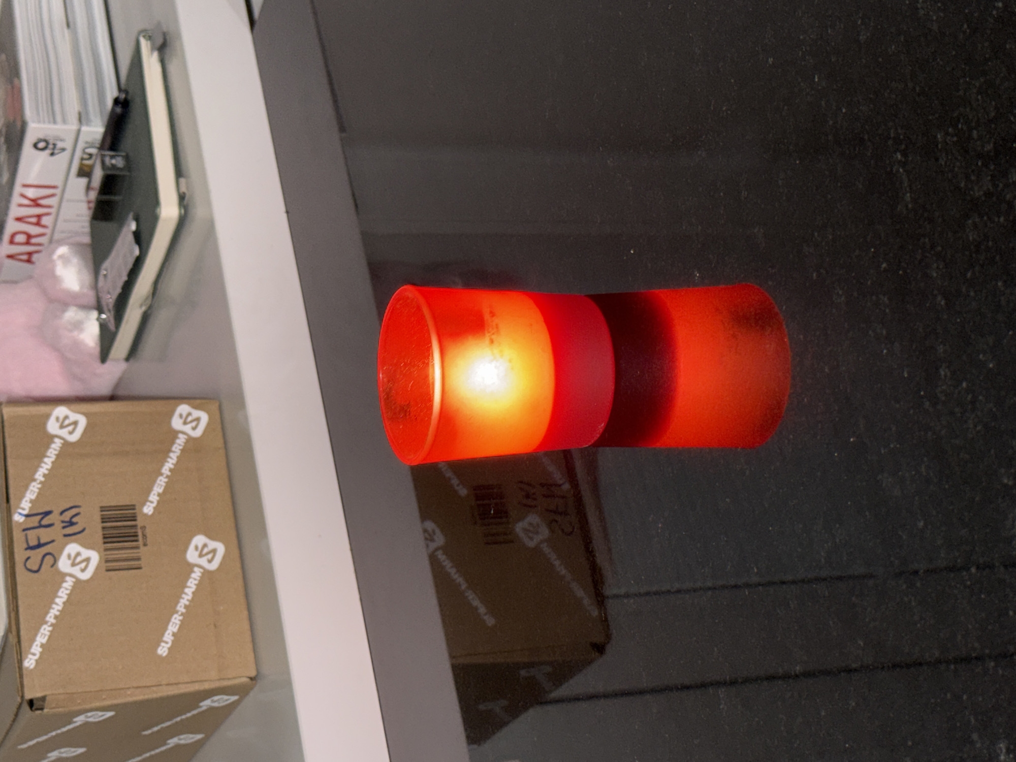 The image shows a close-up of a lit red votive candle on a black reflective surface, likely a countertop. The candle is in a glass holder and emits a warm, orange-yellow light from its flame. In the background, on a white shelf, there is a cardboard box with "SUPER-PHARM" labels and some handwritten text. Next to the box are some books, one of which has "ARAKI" printed on its spine, and a small notebook with a pen. The overall setting appears to be indoors, possibly a domestic or retail environment.