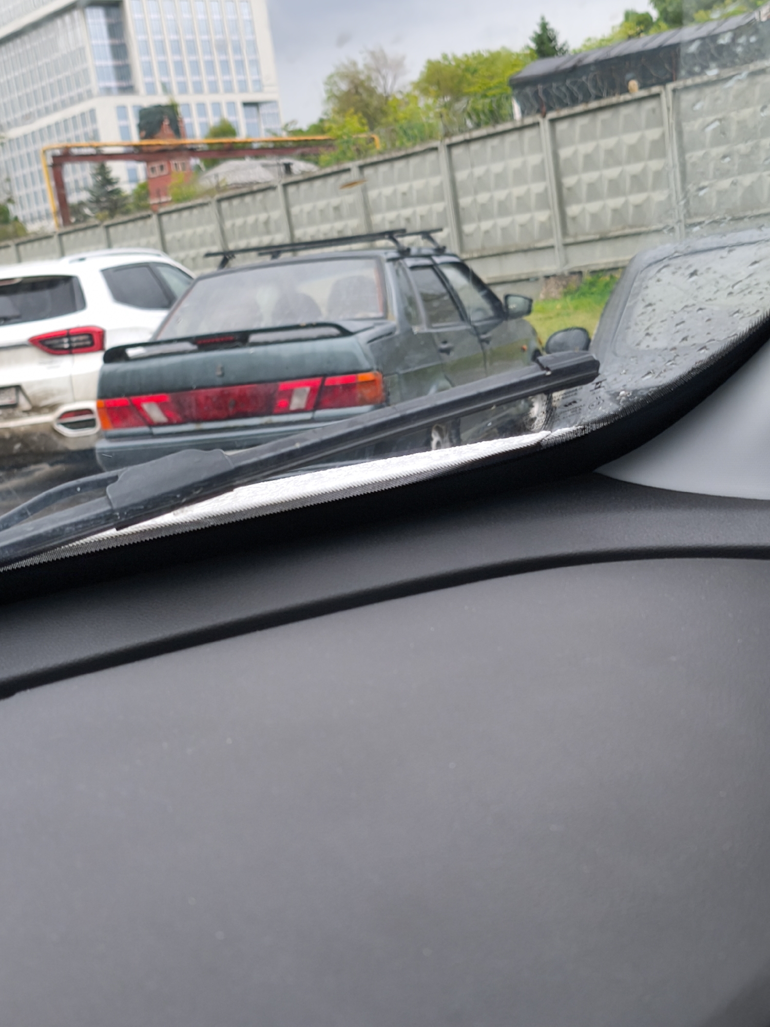 The image shows a view from inside a car, looking out through the windshield. The windshield is wet with raindrops, and a wiper blade is visible. In front of the car, there are two parked vehicles: a dark green car with a roof rack and a white SUV. Behind the cars, there is a concrete fence and a building with a visible industrial pipeline. Given the architecture and infrastructure, the location is most likely within the city of Moscow, Russia.