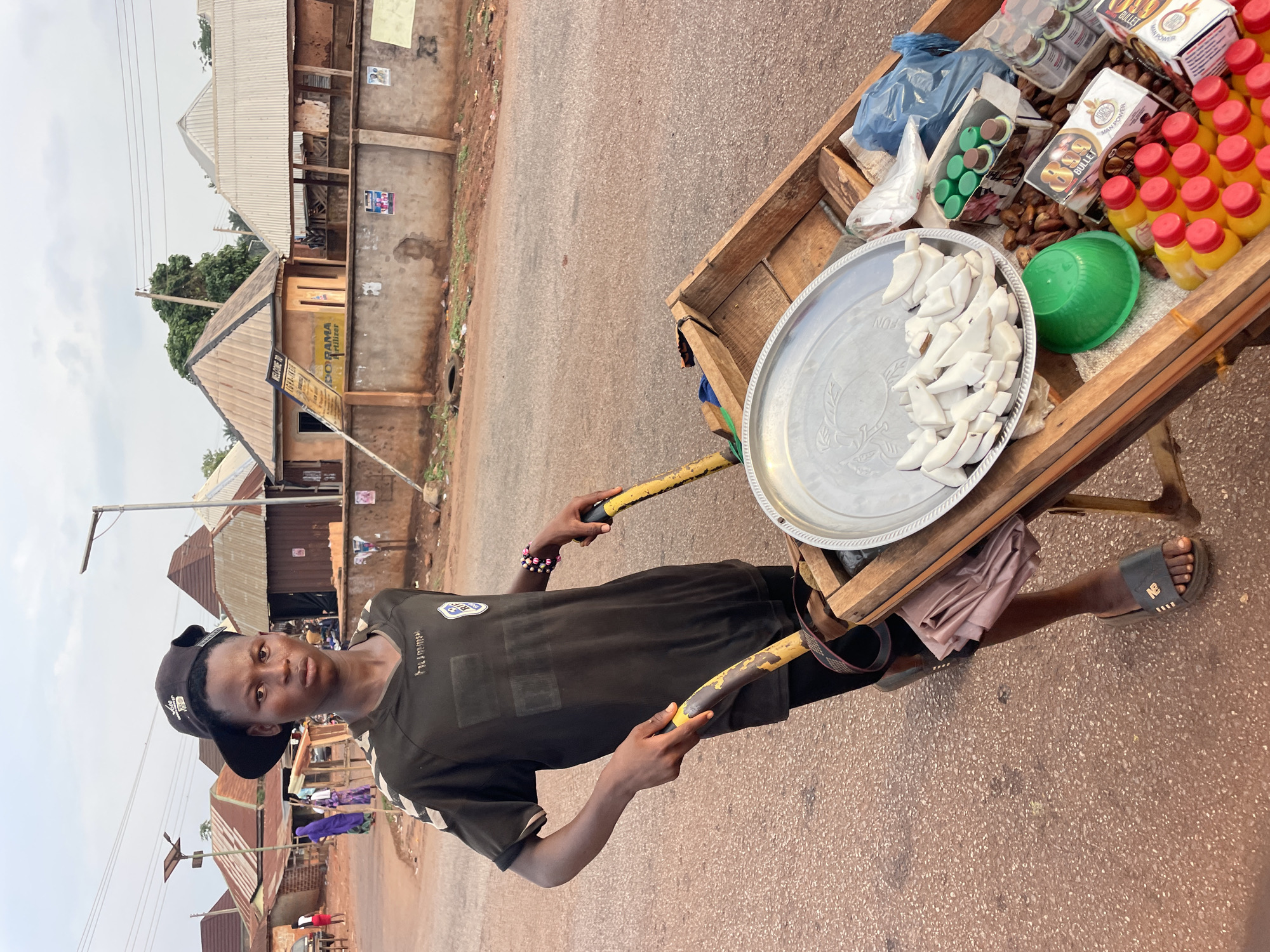 The image shows a young man standing on a red asphalt road in what appears to be Apunu, Nigeria. He is wearing a black t-shirt, black shorts, a black cap and black slippers. He is holding the handles of a wooden cart. The cart is loaded with a silver tray filled with pieces of what appears to be coconut, a green bowl, bottles of orange juice, boxes of what appears to be medicine, and small plastic bags. The background consists of buildings with corrugated metal roofs. A sign is visible on one of the buildings with the words "WELCOME TO IGHA-INEIR DORAMA Fertilizer". Overhead power lines are also visible.