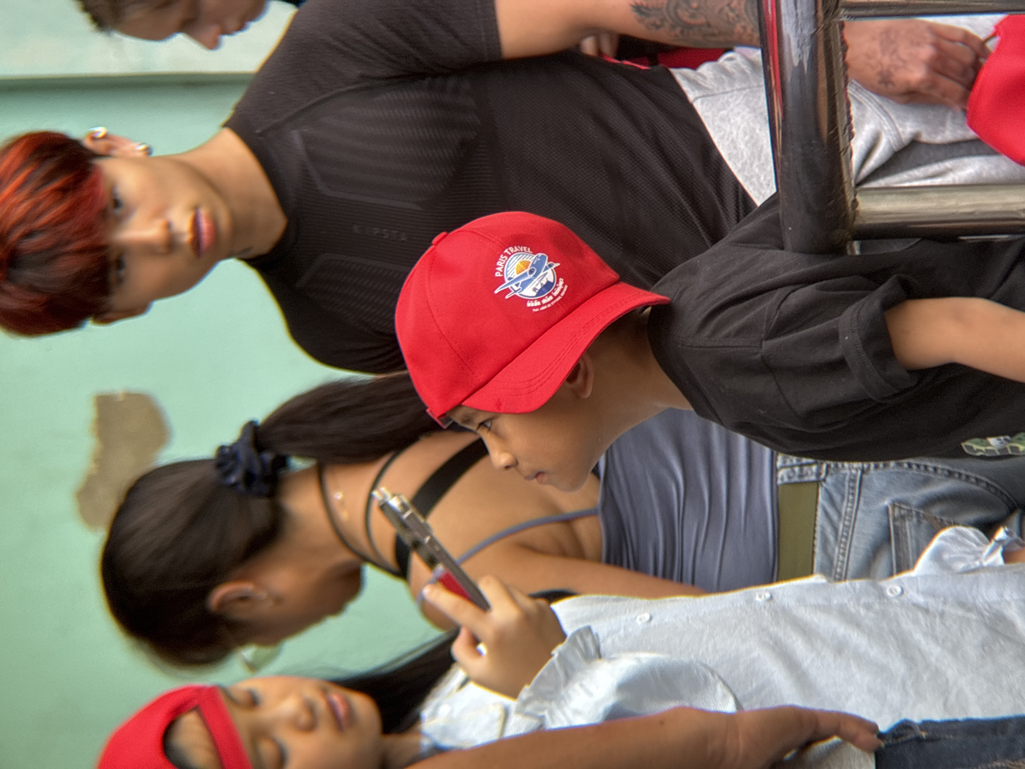 In Lao Bảo Commune, Vietnam, a group of people, including children and young adults, are gathered outdoors. In the foreground, a young boy wearing a red baseball cap with a "Paris Travel" logo on it is looking upwards. Beside him, a young girl in a light blue shirt and jeans is holding a smartphone. Behind them, a young woman with dark hair in a ponytail is wearing a grey tank top and jeans, and a young man with red hair is wearing a black t-shirt. The background shows a light green wall with peeling paint. The scene appears to be a casual gathering or outing.