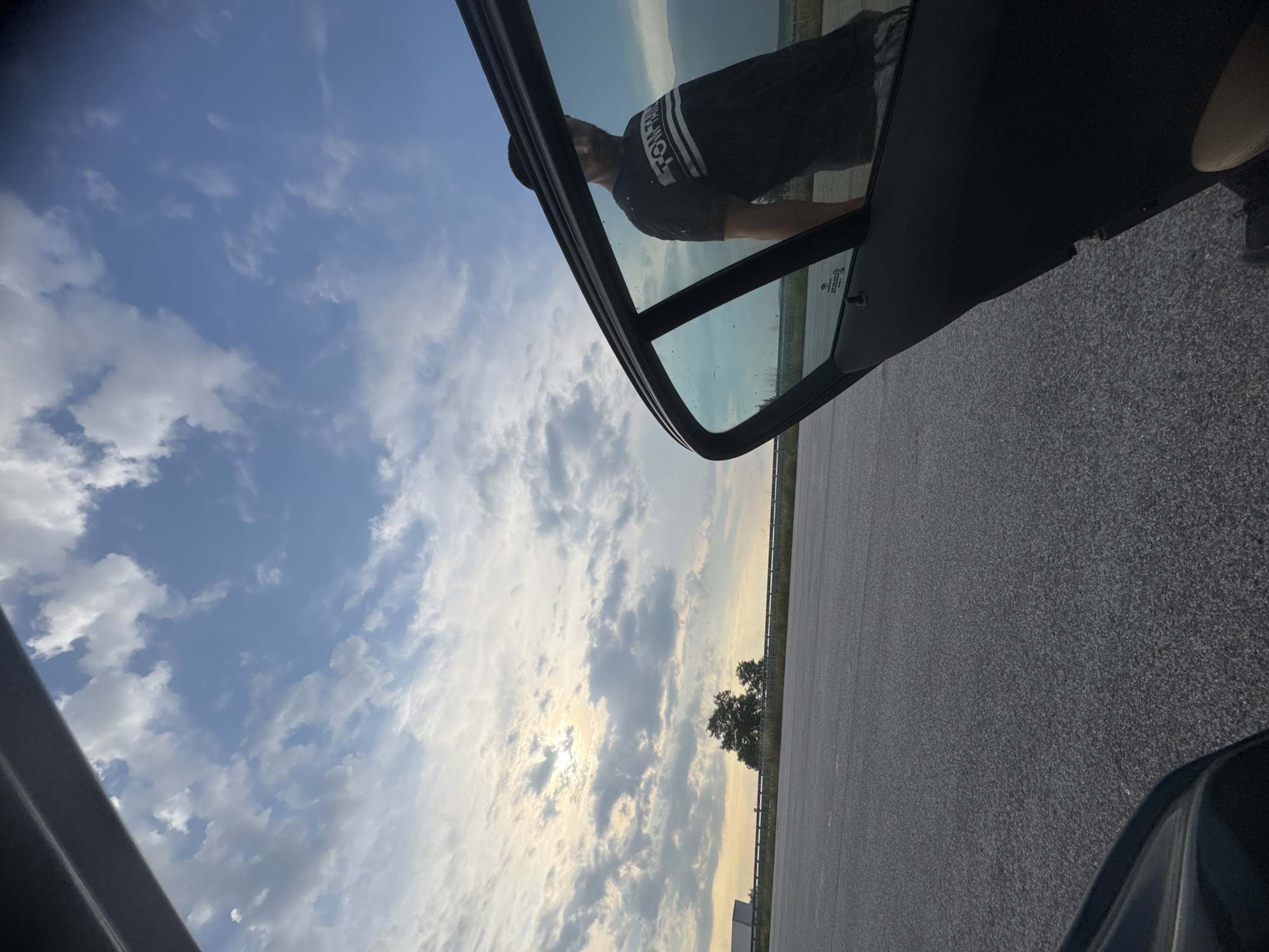 The image depicts an outdoor scene in Rupkite, Bulgaria, at what appears to be a roadside rest area or parking lot during late afternoon or early evening. The upper portion of the frame shows a partially cloudy sky with a bright sun low on the left horizon, casting a strong light through scattered cumulus clouds. The ground consists of dark gray asphalt pavement, extending into the midground. Along the distant horizon, a metal guardrail is visible, with sparse green vegetation and a cluster of trees directly beneath the brightest part of the sky. A large white semi-trailer truck is partially visible on the far left side of the horizon line.
In the foreground and midground on the right, the open front door of a vehicle is prominently displayed, viewed from the car's interior. The car door, including its frame and interior panel, is dark-colored. The door's window is tinted and reflects a male figure and portions of the cloudy sky. The reflected male figure, seen from the upper torso, wears a dark t-shirt with white horizontal stripes and partially legible text that reads "TOM TAN" or similar. His face is somewhat obscured by the reflection and angle, and he appears to be looking towards the left side of the frame. Within the reflection on the car window, a sticker bearing the text "MITSUBISHI MOTORS" is visible on the glass. Faintly visible on the interior window frame is a subtle "BMW" marking. A light tan/beige material, possibly part of the car's interior seating or floor, is visible in the lower right corner.