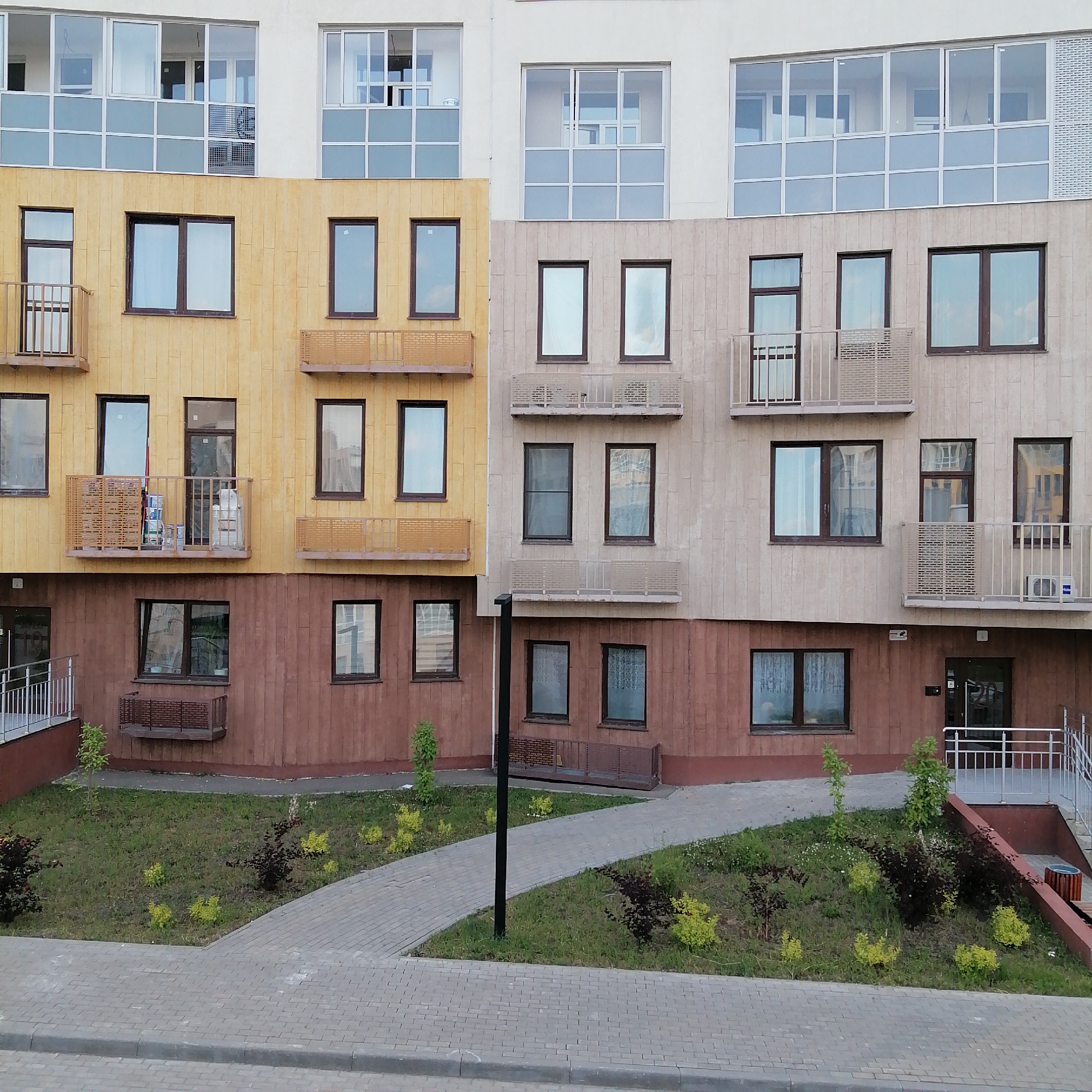 The image shows a modern apartment building in Trubacheyevka, Russia. The building has a multi-colored facade, with sections of yellow, beige, and brown. There are multiple floors with numerous windows and balconies. The ground floor features a landscaped area with grass, small trees, and bushes, along with a paved walkway and steps leading up to an entrance. The walkway is made of gray interlocking pavers. The entrance has a glass door with the number "20" visible. A black lamppost stands near the walkway. The building appears to be new or recently constructed, with some construction materials visible on one of the balconies. The sky is not visible in the frame.