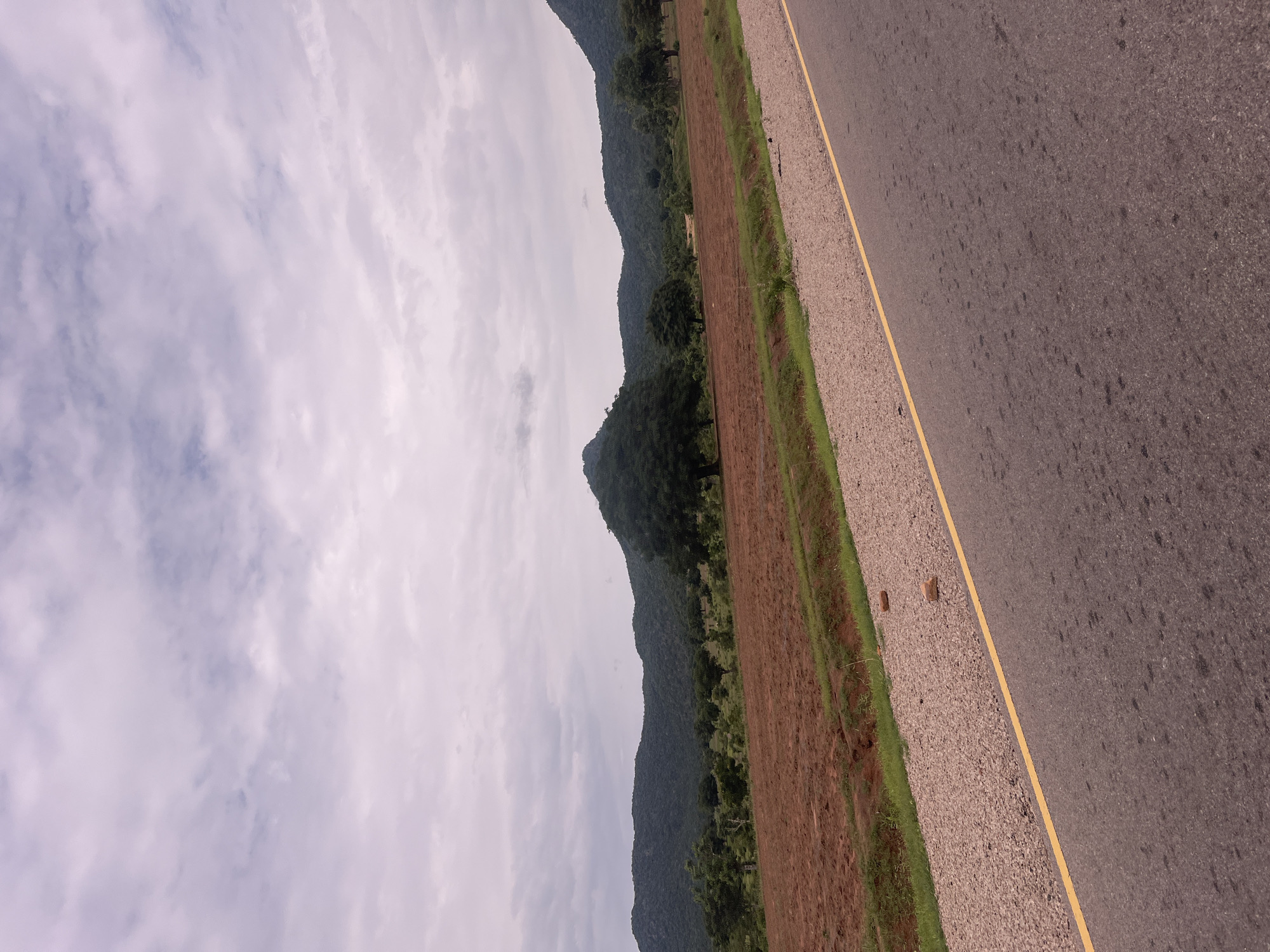 The image depicts a roadside view in Minda, Nigeria. The foreground features a dark asphalt road with a thin yellow line marking the lane. To the left of the road is a gravel shoulder with sparse green grass growing along its edge. Beyond the shoulder, a field of reddish-brown soil stretches, appearing to be recently plowed or prepared for planting. Further in the background, a lush green forested hill rises, covered in dense vegetation and trees. The sky above is overcast with scattered white and grey clouds, suggesting a moderately cloudy day. The lighting is soft, indicating it could be morning or late afternoon. There are no people or discernible objects in the image, focusing instead on the natural landscape and the infrastructure of the road. The scene is peaceful and conveys a sense of rural tranquility.