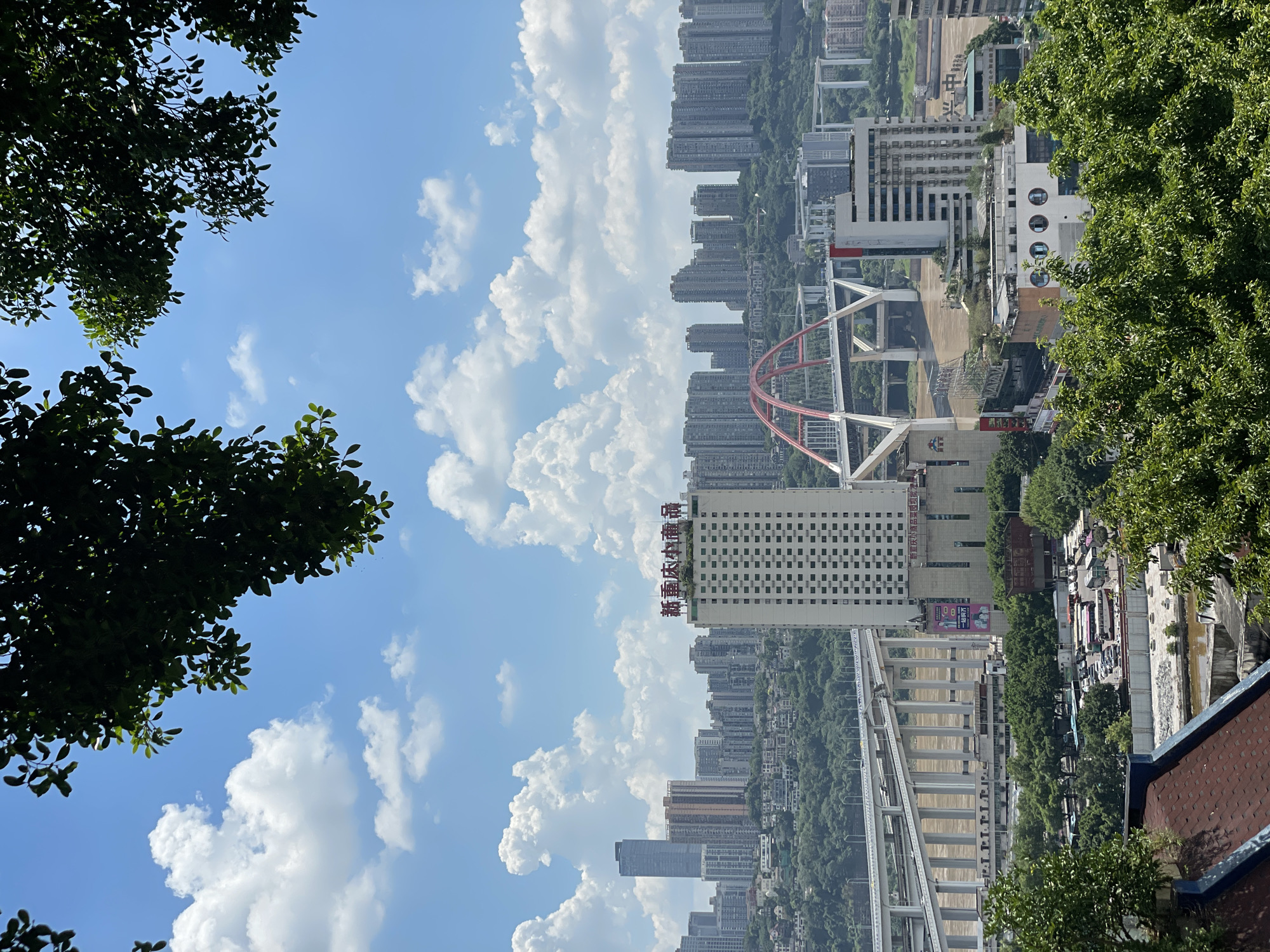 The image captures a panoramic view of the Yuzhong District in Chongqing, China, on a bright, sunny day with scattered clouds. In the foreground, lush green trees frame the scene from the top and bottom edges. A significant portion of the image is dominated by the sprawling cityscape, characterized by numerous tall buildings and high-rise apartments that ascend hillsides.
A prominent feature is a large, multi-tiered highway bridge crossing a wide, brown river, likely the Yangtze or Jialing River, which flows through the city. To the right of the bridge, a distinctive red-arched bridge adds a striking architectural element to the urban landscape. A tall, white rectangular building with a grid-like facade and Chinese characters on its top stands out among the surrounding structures. Below the bridges and buildings, the scene transitions to a more densely populated area with smaller buildings, roads, and a parking lot filled with various vehicles including buses and cars.
The overall impression is one of a vibrant, modern metropolis situated amidst a hilly terrain, with extensive infrastructure and a dynamic urban environment. The sky is a vivid blue, dotted with fluffy white cumulus clouds, contributing to the overall bright and clear atmosphere of the scene. No people are clearly discernible in the image.