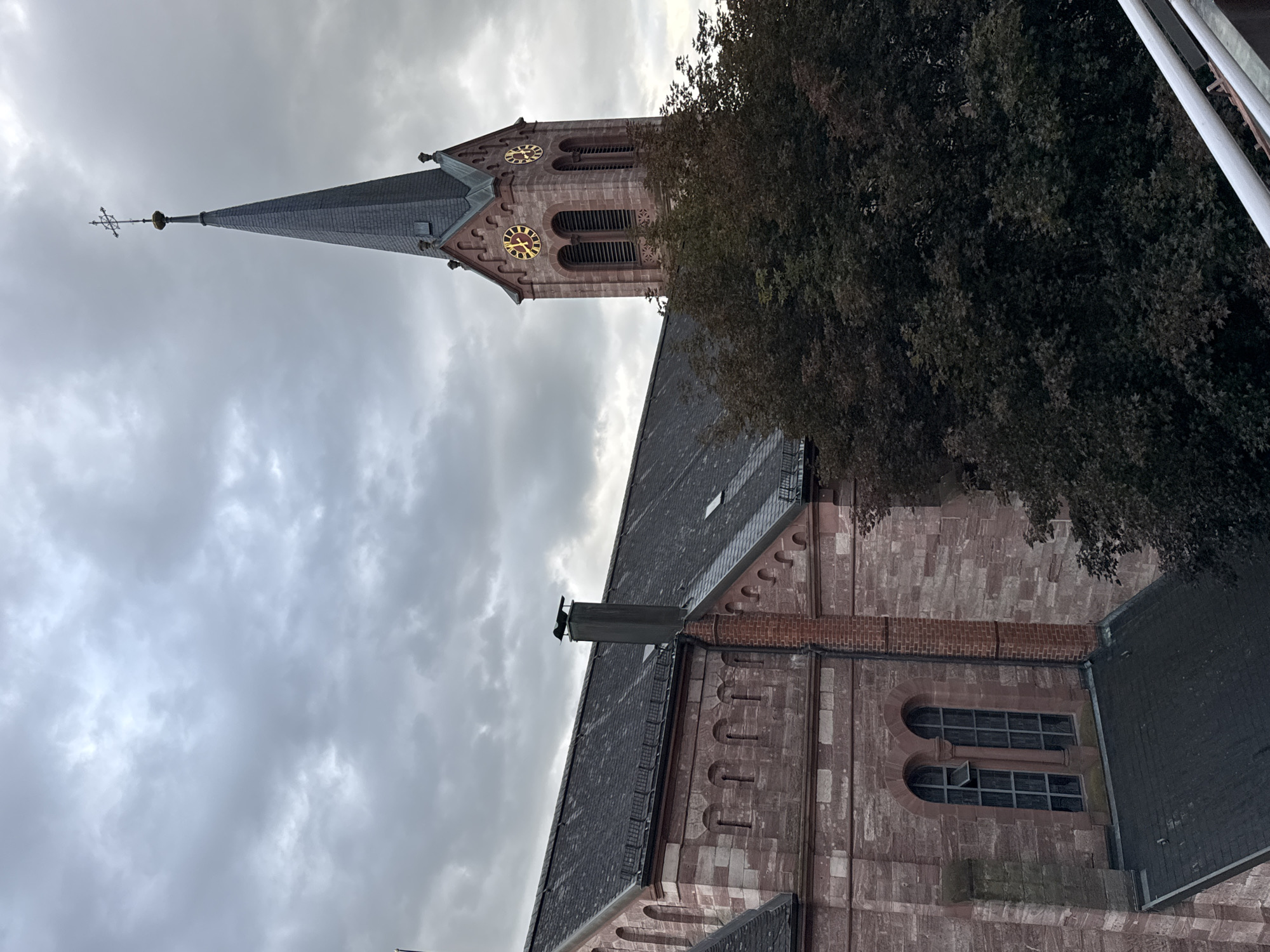 This is a medium-distance, eye-level shot of the Protestant Church of Schopfheim, Germany, under a cloudy sky. The church, constructed with reddish-brown stone, features a tall, dark-shingled spire topped with a cross. Two clock faces are visible on the tower, with Roman numerals. The building's roof is also shingled. To the right of the church tower, a dense canopy of dark green trees obscures a portion of the building. A brick chimney with a black decorative cap is visible on the roof of the main church structure. The image composition focuses on the church and its surroundings, with no people present.
