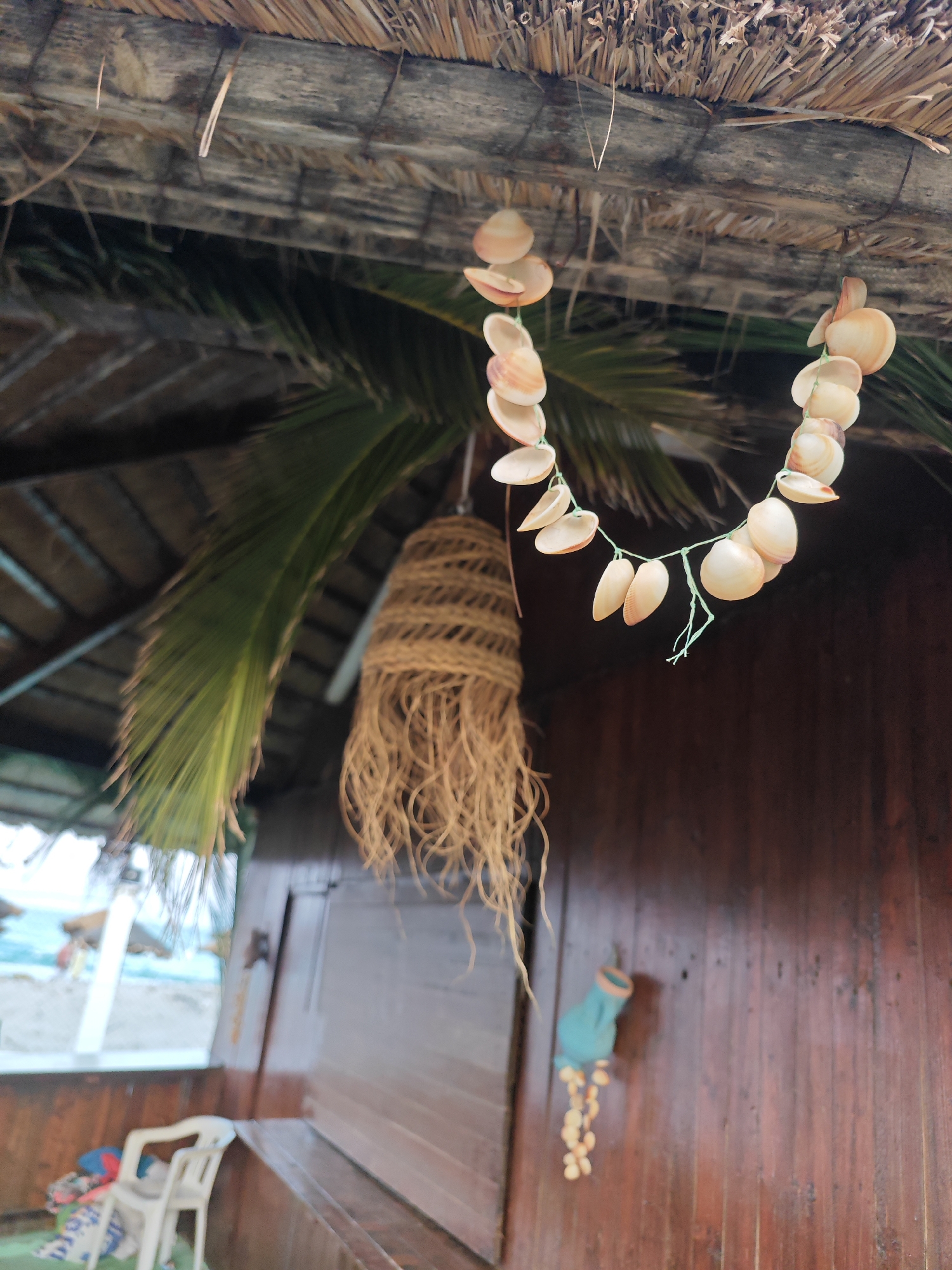 This is a close-up, medium shot of a thatched roof overhang with hanging decorations. Two strings of seashells, light beige and pinkish-orange, are strung across the scene. To the left, a palm frond hangs down, obscuring part of the background. Beneath the palm frond, a hanging object made of twine with frayed ends dangles. The background consists of a dark brown wooden wall and a blurred view through an opening to a beach with turquoise water and a few indistinct figures. A white plastic chair and some colorful items are visible in the lower left corner. The scene suggests an outdoor, possibly seaside, location in Hiboun, Tunisia.