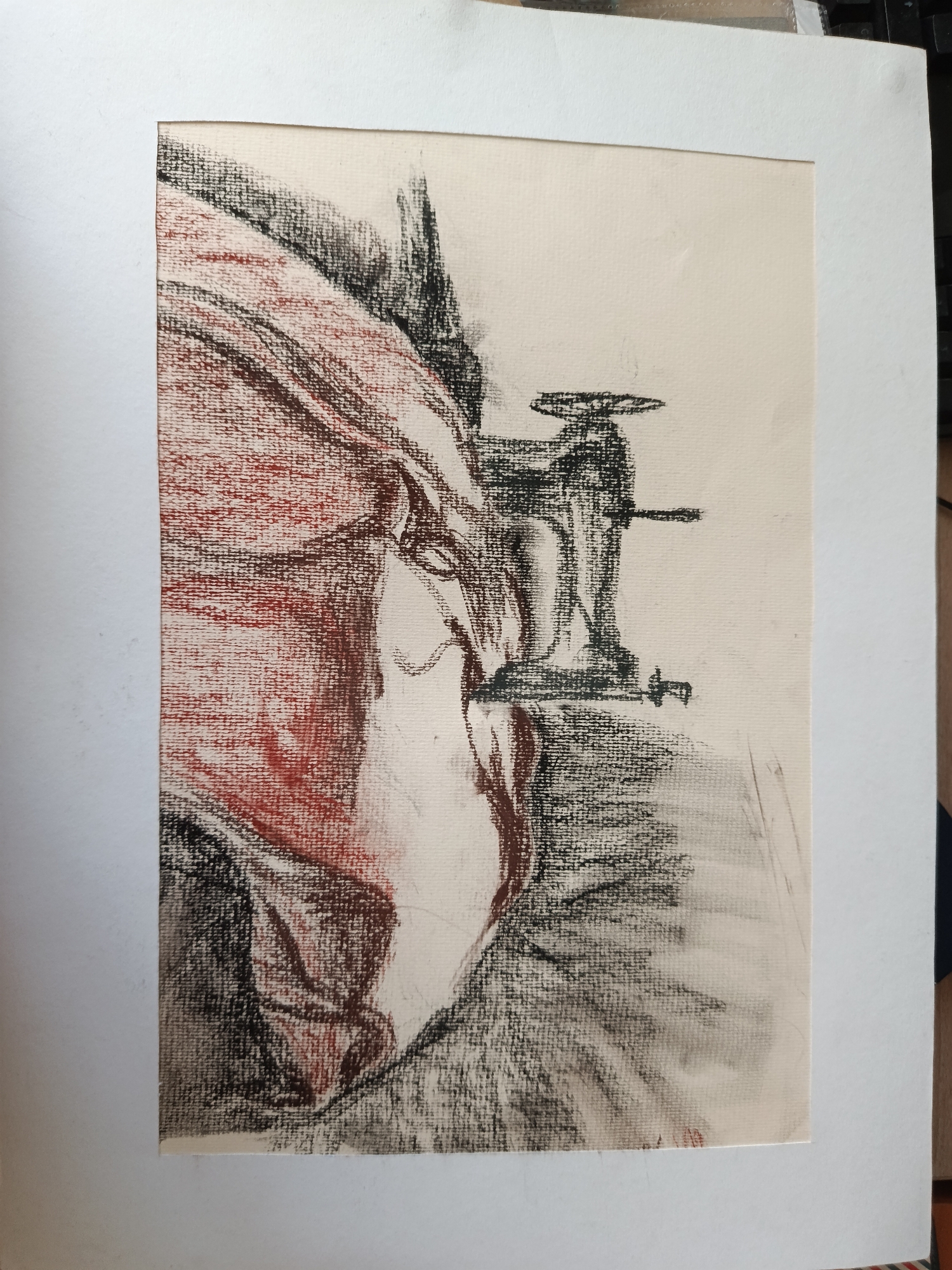 A charcoal and chalk drawing on paper, matted in white, depicts drapery and a vise. The drapery is primarily rendered in red chalk and charcoal, with lighter areas suggesting folds and highlights. The vise, positioned to the right of the drapery, is drawn in black charcoal and features a handle, screw, and clamping jaws. The background is off-white, providing contrast to the dark charcoal and chalk. The location of the drawing is not discernible.