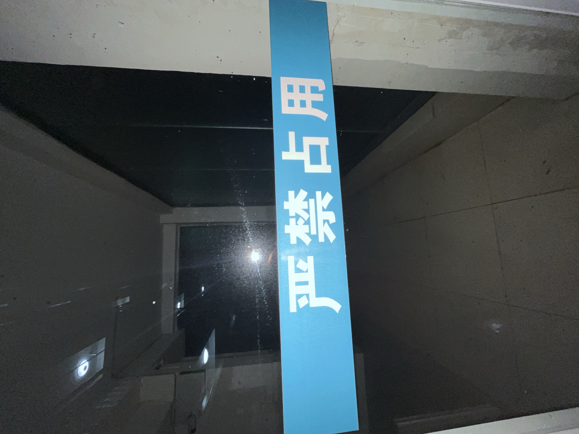 The image depicts an indoor scene focused on a translucent glass partition or door, likely located in Yuzhong District, China. A prominent horizontal blue strip is affixed to the glass at eye level, displaying four white Chinese characters: "严禁占用" (Yánjìn zhànyòng), which translates to "Strictly forbidden to occupy" or "No occupying."
Through the glass, a dimly lit interior corridor or hallway is visible. The floor appears to be a light-colored, tiled surface. Overhead, bright, circular recessed light fixtures are reflected in the glass, indicating an illuminated ceiling above the vantage point. The area beyond the glass seems darker, with a dark ceiling and walls, possibly also made of glass or dark paneling. Multiple reflections and some glare are present on the glass surface, suggesting internal light sources. The glass itself exhibits some smudges and dust, particularly visible around the reflections.
To the right of the glass partition, a vertical, light-colored structural column or wall frame is partially visible, showing some minor wear or discoloration. No individuals or specific objects other than the architectural elements and lighting are discernible. The scene presents a utilitarian, institutional aesthetic.
