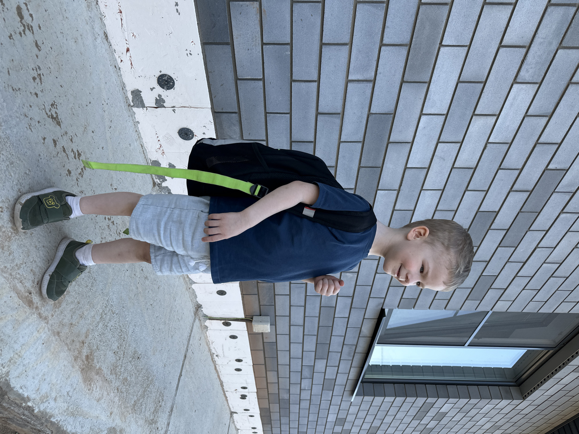 A young boy stands on a concrete walkway next to a brick building. He is wearing a dark blue t-shirt, light gray shorts, white socks, and olive green shoes with a bear emblem. He has a black backpack with a bright green strap over his left shoulder. The building is made of gray bricks arranged in a running bond pattern. A window is visible to the right of the boy. The setting appears to be an urban or suburban environment. It's not possible to confirm the specific location in Прикамский, Russia, without additional visual or contextual clues.