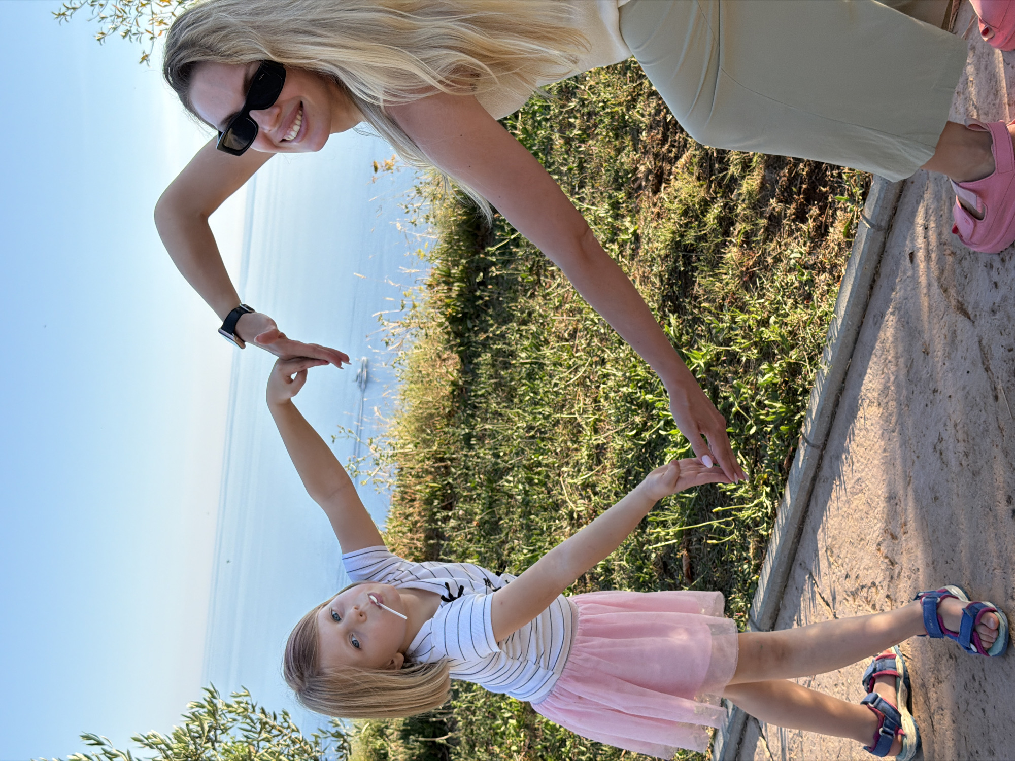 The image shows two people, a woman and a young girl, in an outdoor setting with a body of water in the background. The woman is on the left, wearing sunglasses and smiling. She has blonde hair and is wearing a light-colored top and light-colored pants. Her left arm is extended, and her hand is open. The girl is on the right, facing the camera and holding a lollipop in her mouth. She has blonde hair and is wearing a striped t-shirt and a pink tutu skirt. Her arms are extended, and her hands are reaching out towards the woman's hand, forming a heart shape with their fingers. The scene appears to be at Kovačko Polje, Montenegro. The sun is shining, suggesting it is daytime. The overall mood seems joyful and playful.