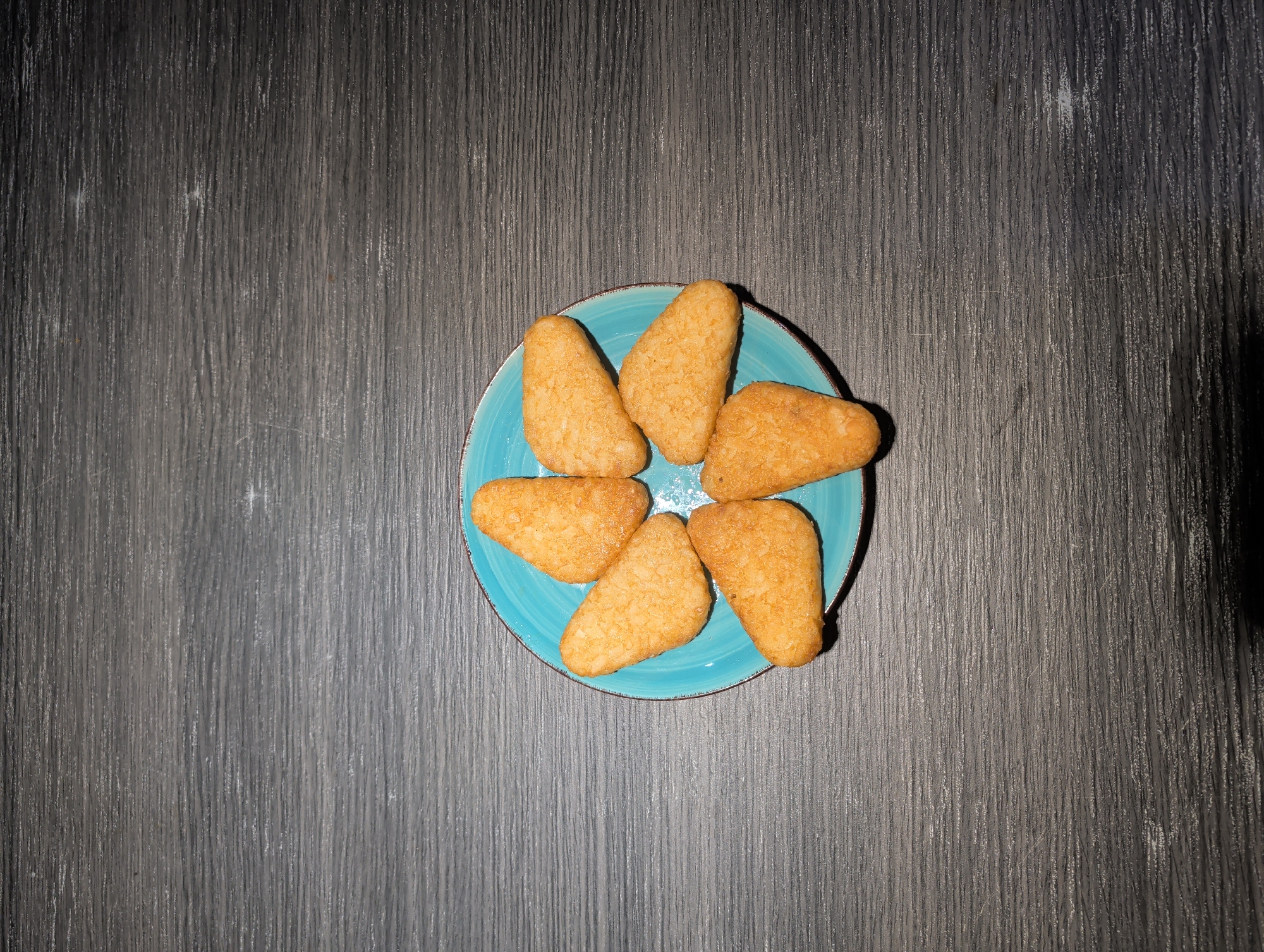 A top-down, full shot shows seven hash browns arranged in a circle on a turquoise plate with a brown rim. The plate is centered on a gray, wood-grain textured surface. The lighting is brighter on the left side of the image, casting a shadow on the right. There are no people visible. The location is not determinable from the image.