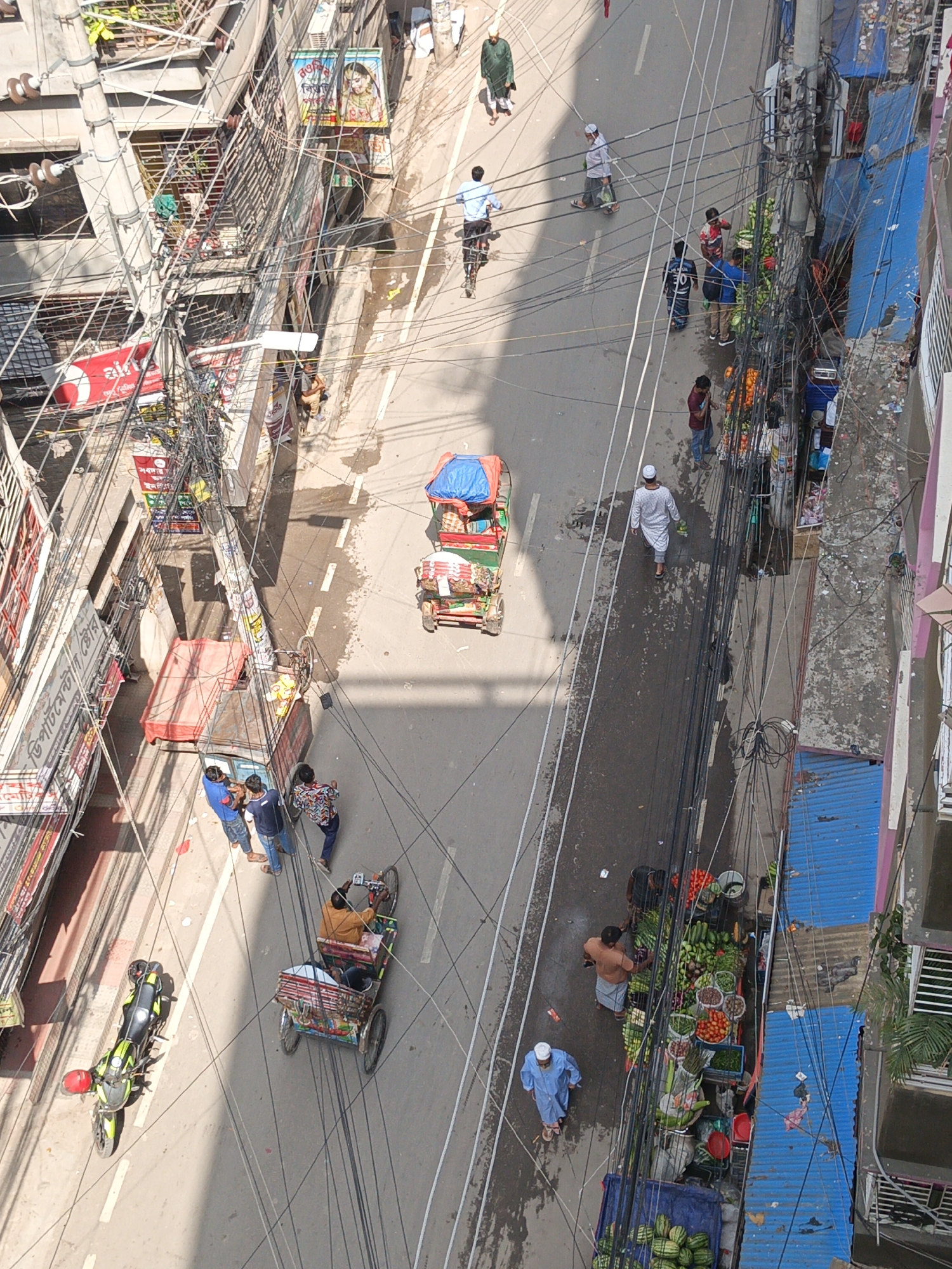 An aerial view of a street in Dhaka, Bangladesh shows various activities. Numerous power lines run across the street, obscuring some details.
People are walking, cycling, and engaged in commerce. Several rickshaws and hand-pulled carts are on the street. A fruit and vegetable vendor is selling their wares on the right side of the street. Some people appear to be making purchases. A motorcycle is parked on the left side of the street. Buildings line both sides of the street, with signs and awnings visible.