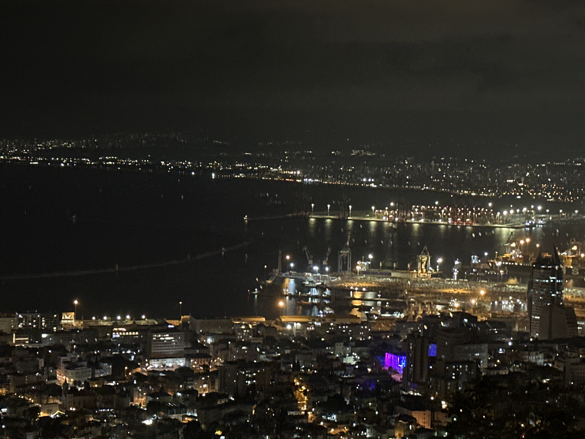 This is a nighttime aerial photograph of the city of Haifa, Israel. The foreground displays a densely populated urban area with numerous buildings illuminated by interior lights. In the midground, a harbor is visible, featuring a collection of cranes, cargo ships, and docks illuminated by port lights. Beyond the harbor, the cityscape of Haifa stretches towards the horizon, with lights from buildings and streetlights creating a sprawling network of illumination. The background is dominated by the dark expanse of the sea, with distant lights suggesting further urban development along the coastline. The sky is dark and overcast.