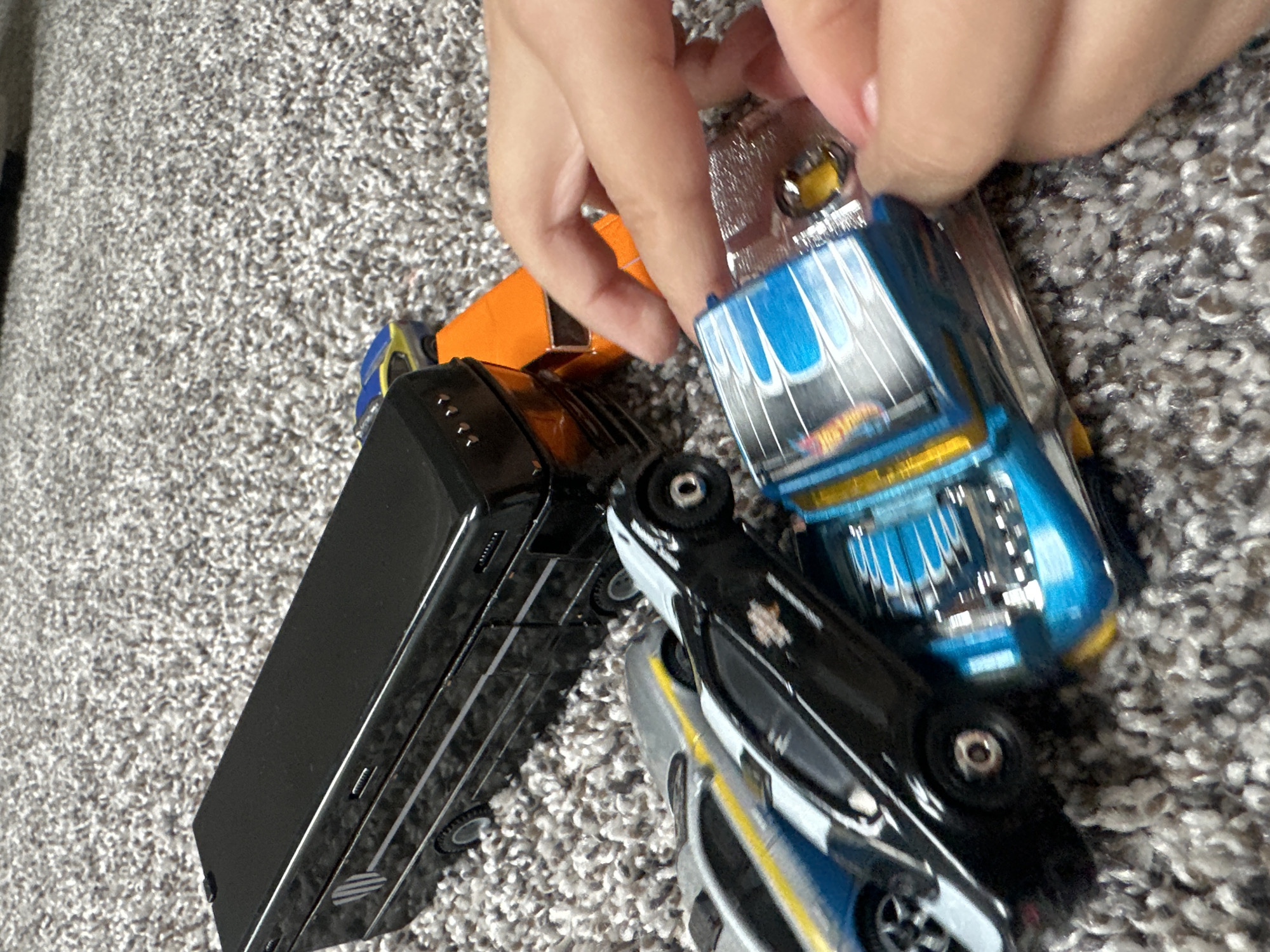 A close-up, eye-level, indoor shot depicts a collection of toy cars scattered on a grey, textured carpet, presumably in Schaumburg, United States of America. A black bus-like toy vehicle is positioned in the upper left of the frame, with its side facing the viewer. To the right of the black bus, an orange toy car is visible. A silver and blue toy car with a yellow stripe is partially obscured by other vehicles. A black toy car with white markings is in the foreground, angled towards the viewer. A blue and silver toy car with flame graphics is the most prominent object in the foreground, being handled by a child's hand with painted fingernails. The child's fingers are touching the chrome rear of the blue and silver car. The scene is brightly lit, suggesting it was taken during the day.