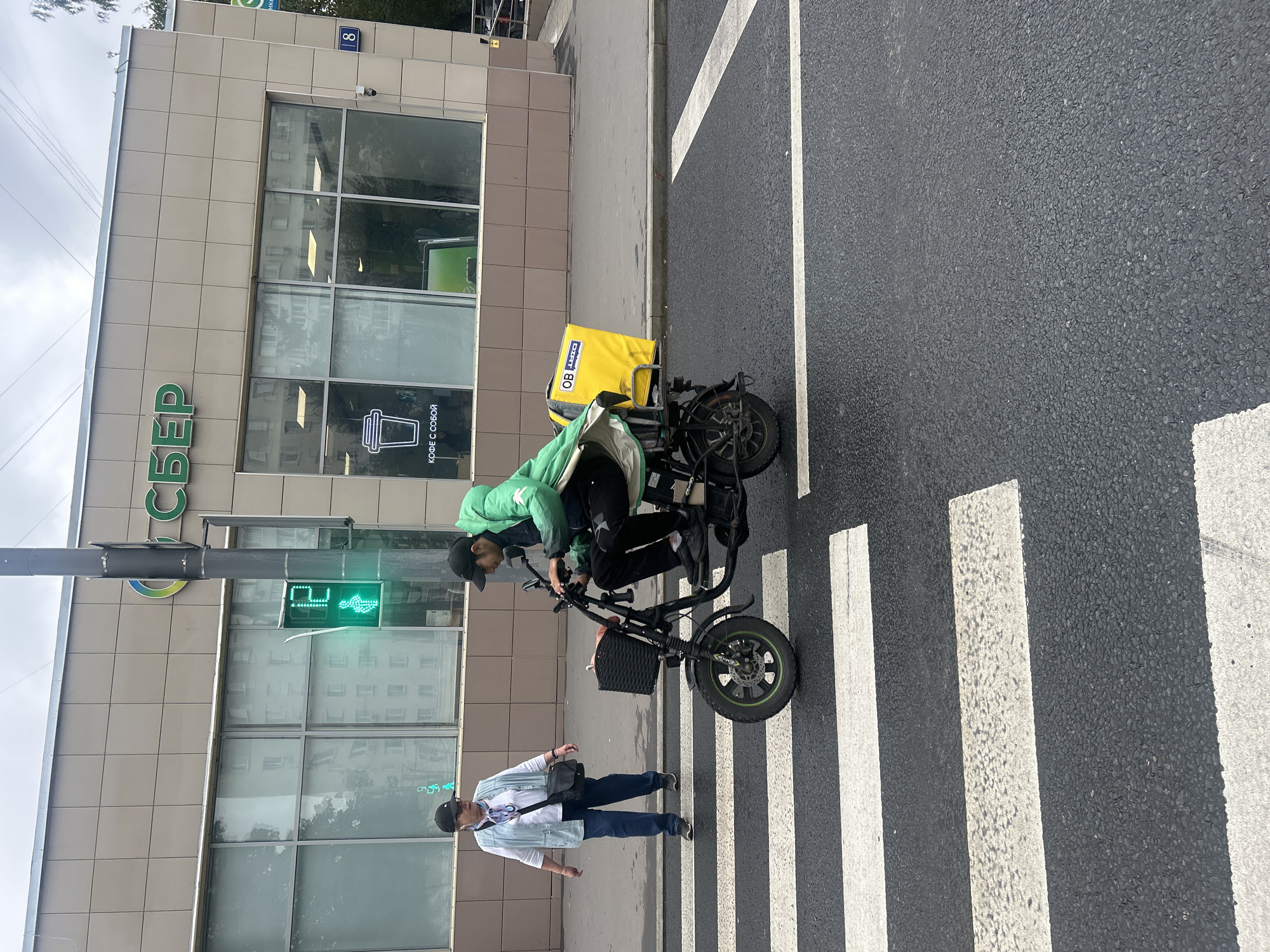 In Moscow, Russia, on a cloudy day, a man wearing a green jacket and a black cap rides a black electric scooter with a large yellow delivery box on the back. He is on a crosswalk, with a traffic light showing a green walking figure. To his left, a woman in a light blue vest, white shirt, and dark jeans stands on the crosswalk, facing the scooter. Behind them is a building with a large sign that reads "СБЕР" and a window displaying a coffee cup logo.