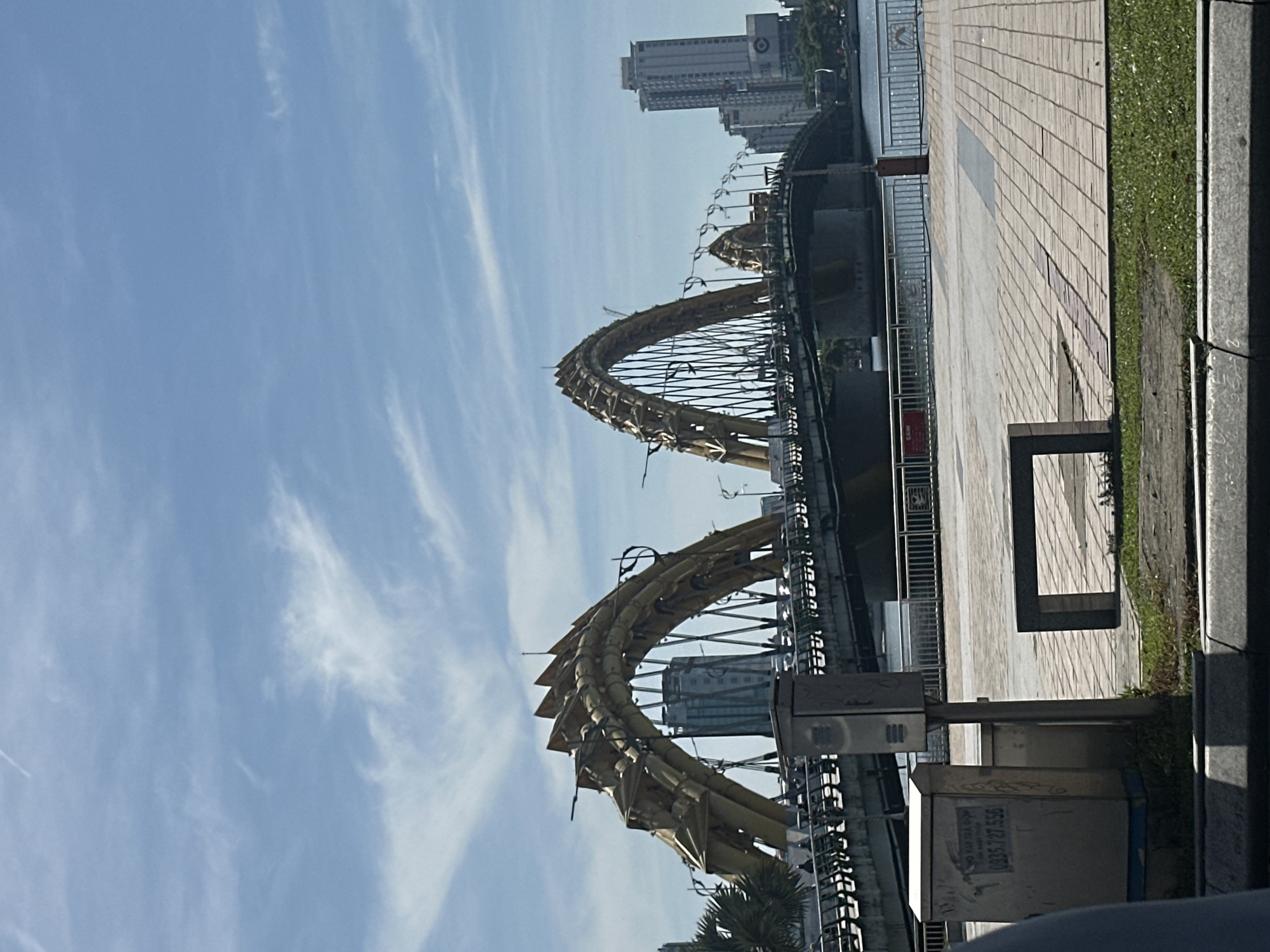 The image shows the Dragon Bridge in Da Nang, Vietnam, on a clear day. The bridge, a prominent landmark, is designed to resemble a dragon and spans a body of water. In the background, modern buildings and a palm tree are visible. The foreground features a paved walkway, a bench, a metal box with some writing on it, and grass. The sky is blue with scattered white clouds.