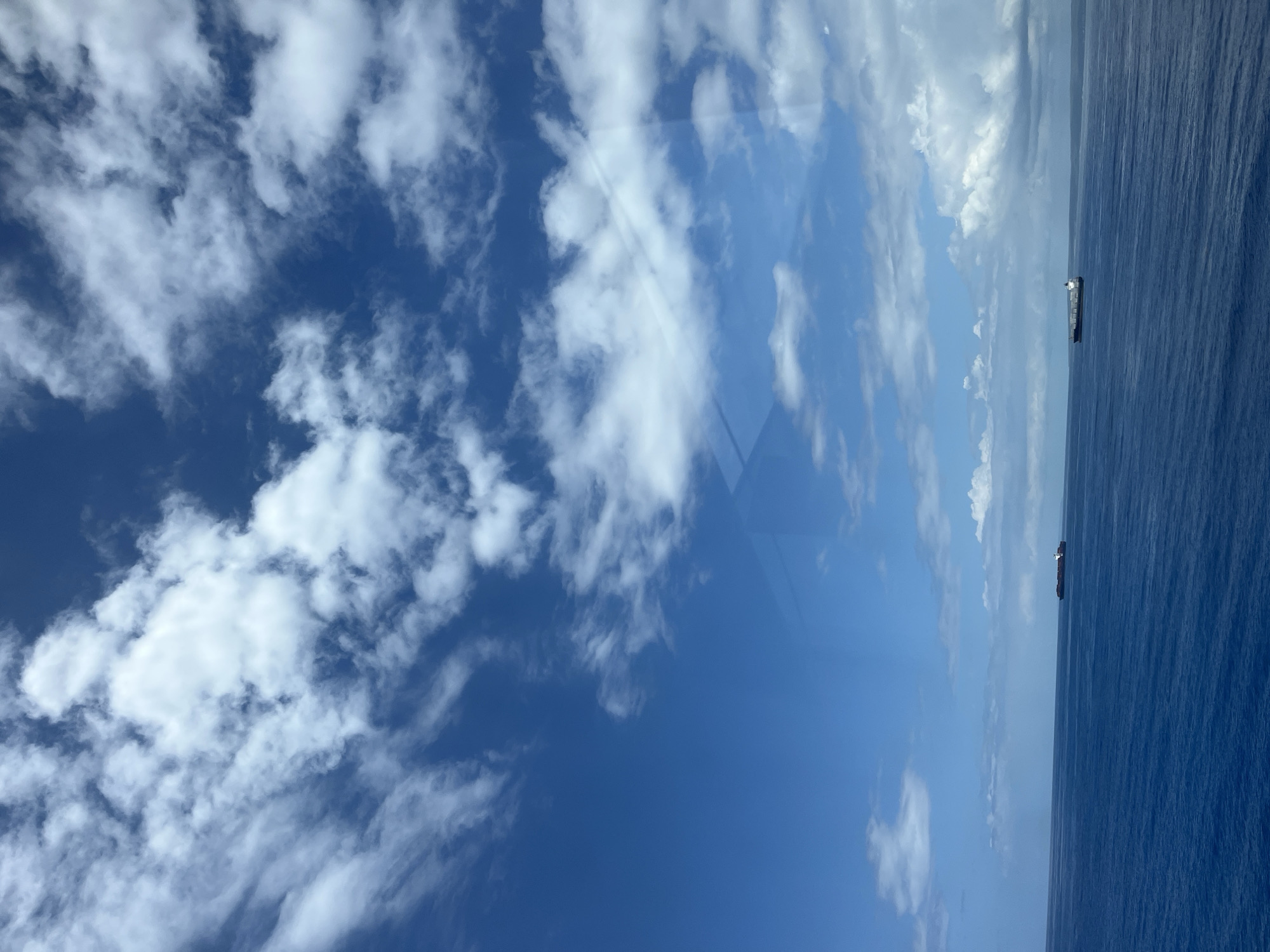 The image shows a view of the ocean and sky, with a few clouds scattered across a deep blue sky. In the distance, two ships are visible on the horizon line. The image is taken from an elevated perspective, possibly from a building or higher ground. The image does not provide any details about the location or the presence of people.