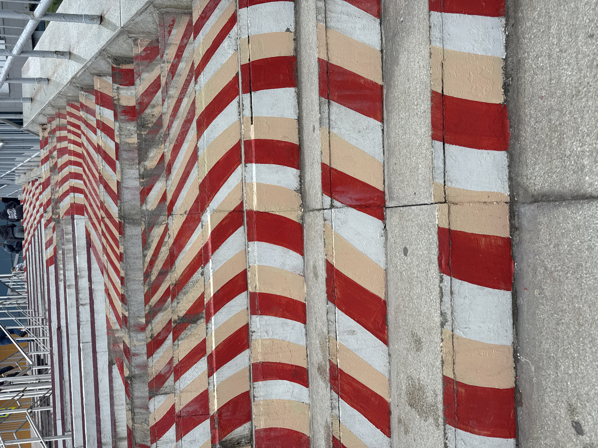 The image shows a wide outdoor staircase with a distinctive red and white striped pattern painted on the risers. The stripes are angled and alternate between red and a lighter color, possibly beige or a faded white. The stairs are made of concrete and appear to be well-worn. In the background, people are visible walking up the stairs. One person is wearing a dark hooded sweatshirt, and another is wearing blue jeans. The staircase has metal handrails on the sides. The environment appears to be urban, with hints of buildings and more stairs in the distance. The lighting suggests it is daytime. This staircase is located in Ufa, Russia, specifically at the Monument to Salavat Yulaev.