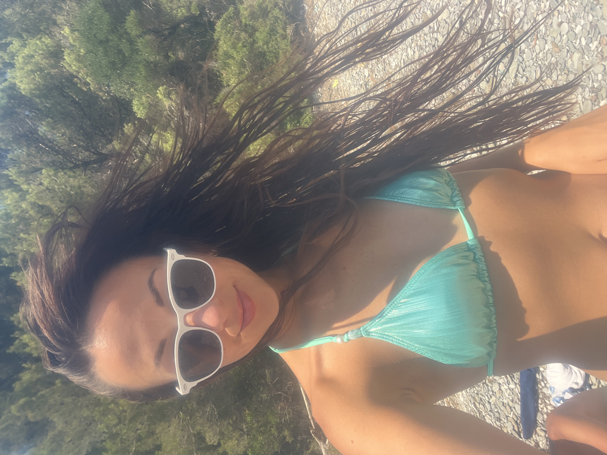 The media file shows a close-up, overhead shot of a woman wearing a turquoise bikini and white sunglasses. She has long, dark, wet hair that is blowing in the wind. Her skin is tanned and appears to be illuminated by the sun. The background consists of green foliage and a rocky, pebbly ground. The lighting suggests it is daytime, likely a sunny day. The scene appears to be outdoors, possibly at a beach or a scenic viewpoint, given the natural elements visible. The woman's face is partially visible, and she is smiling. The overall impression is one of a relaxed, possibly vacation setting. There is no visible text in the image.