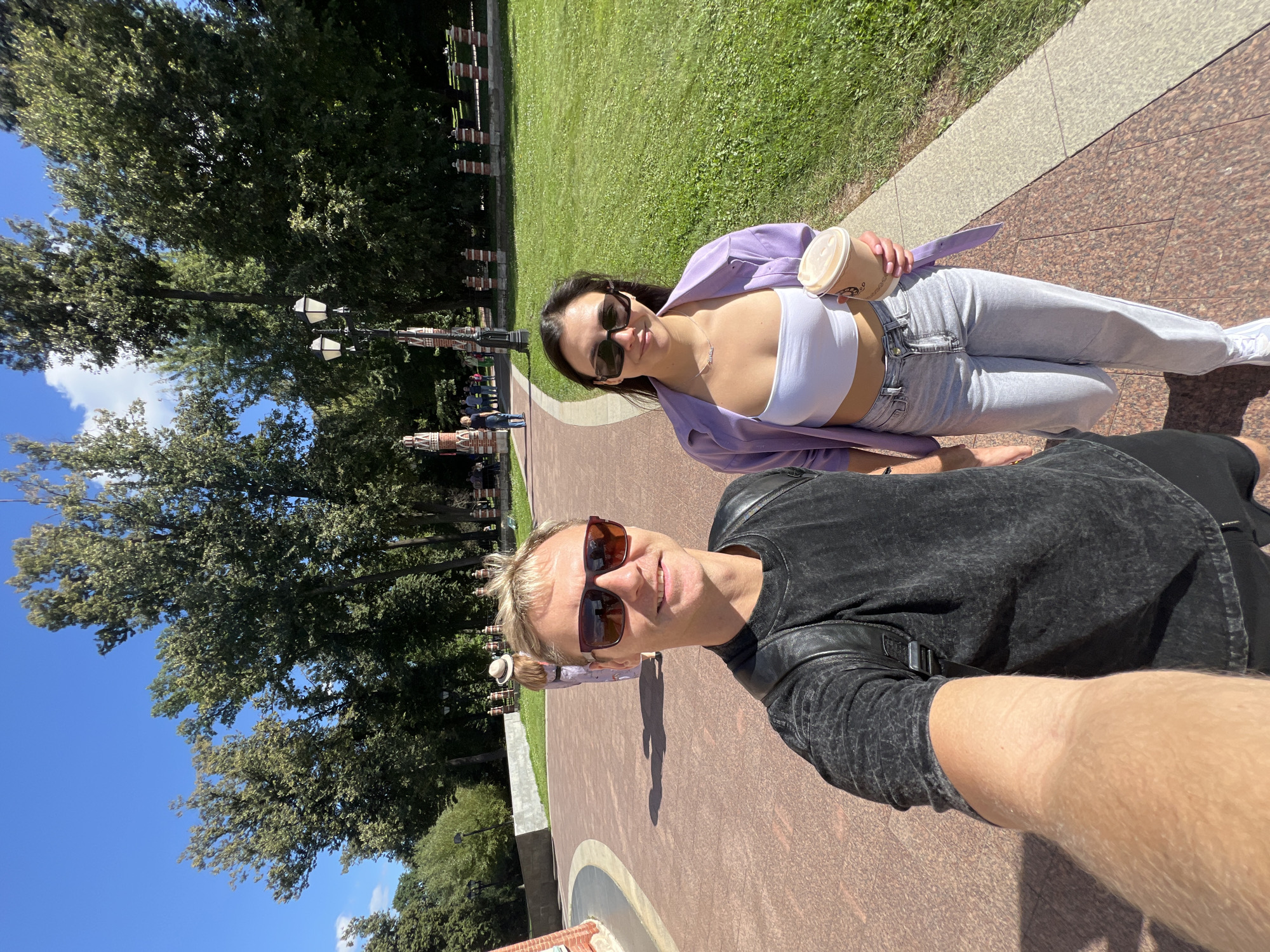 Two individuals, a man and a woman, are posing for a selfie in a park on a sunny day. The man, in the foreground, is wearing a dark, distressed t-shirt and sunglasses. The woman, standing beside him, is wearing a white crop top, a light purple unbuttoned shirt, and light wash jeans. She is holding a to-go cup. They are on a paved walkway in what appears to be a park with trees and grassy areas. In the background, other people are visible walking along the path, and there are decorative brick structures and lampposts. The environment suggests a public park or garden setting. The location is identified as Tsaritsyno Park in Moscow, Russia.